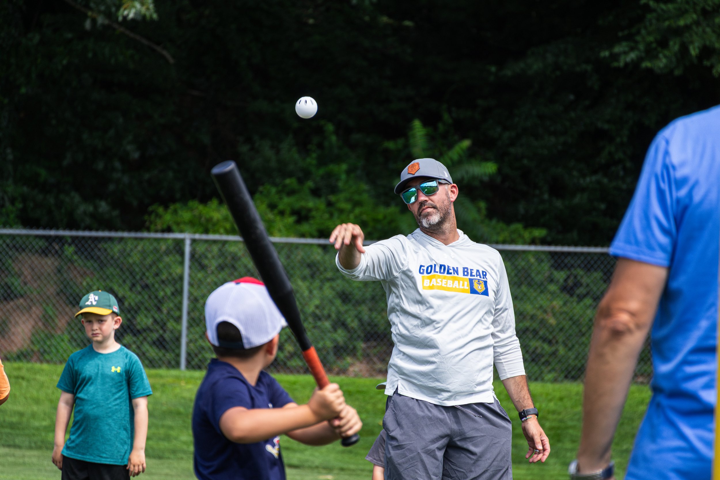 A man wearing sunglasses, a cap, and a white Golden Bear Baseball shirt is coaching kids during a baseball practice on a grassy field with a chain-link fence and trees in the background.