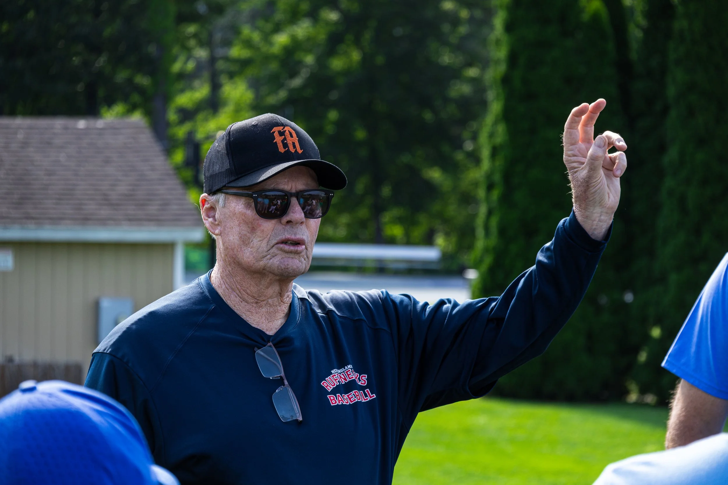 An elderly man wearing sunglasses, a black cap with an orange logo, and a navy blue shirt with red and white lettering, is raising his right hand outdoors during daytime, possibly speaking or giving instructions.
