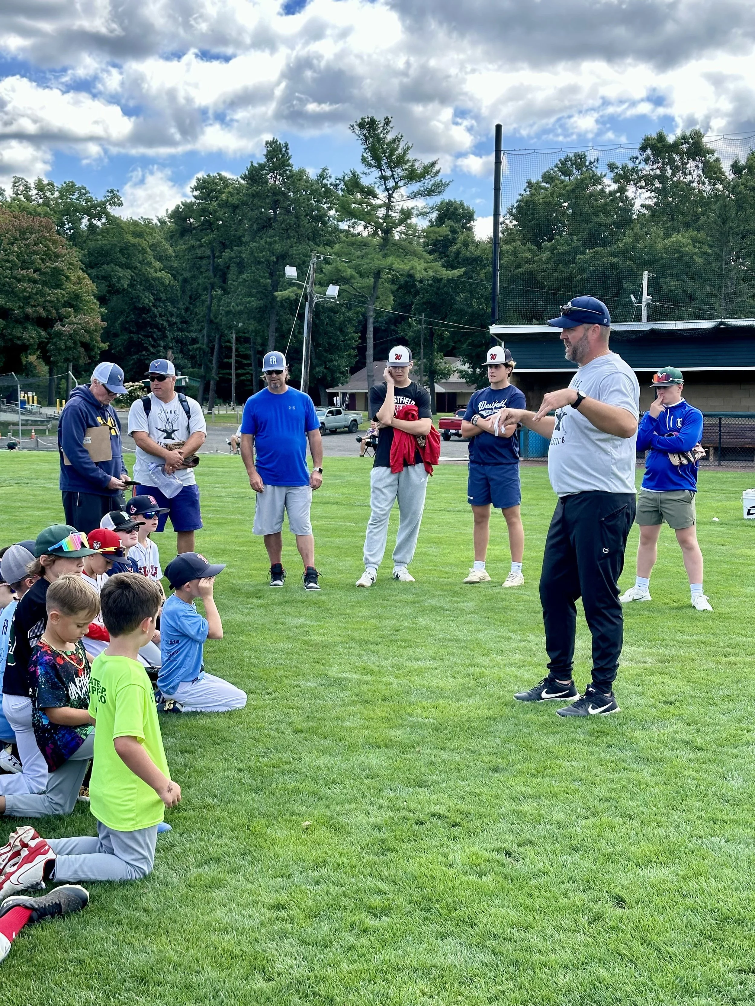 A coach speaking to a group of young baseball players kneeling on the grass at a baseball field during daytime. Several adults and players wearing caps and sportswear are present, with tall trees and a cloudy sky in the background.