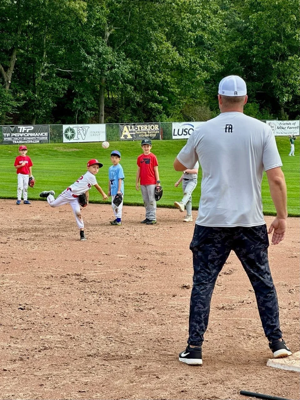 A group of young boys on a baseball field during practice, with one boy in a white uniform throwing a ball, and a coach or instructor standing in front of them, facing away.