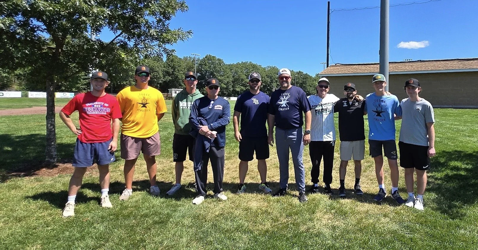 Group of ten men standing outdoors on grass with trees and a building in the background, dressed in casual athletic clothing, some wearing baseball caps, smiling at the camera.