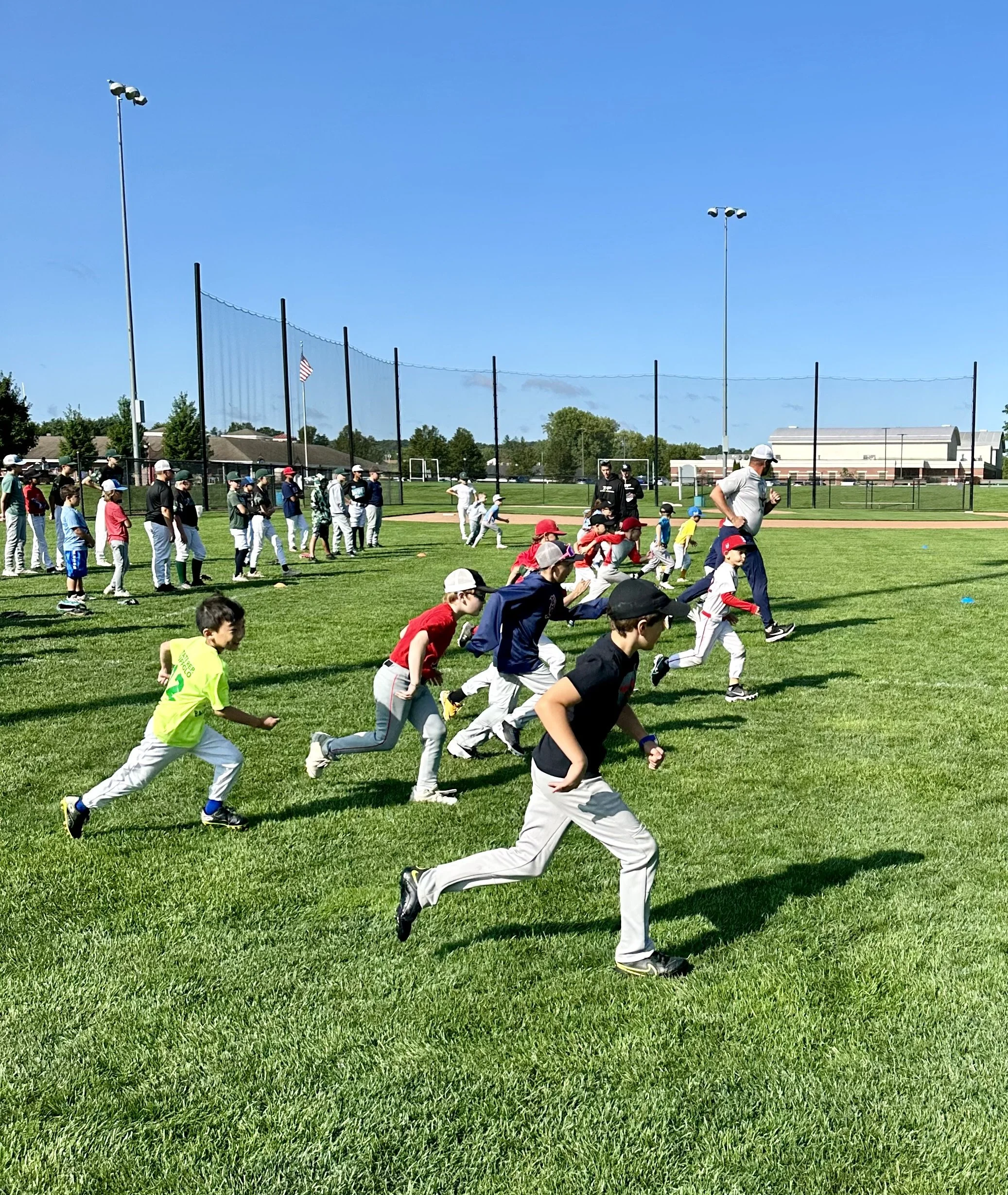 Children running and playing on a baseball field during daytime under blue skies with a tall light pole and a chain link fence in the background.