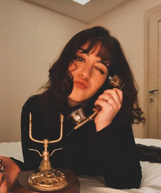 Young woman with dark curly hair making a kiss face, holding a vintage telephone receiver to her ear, sitting on a bed with a brass candelabrum in front of her.