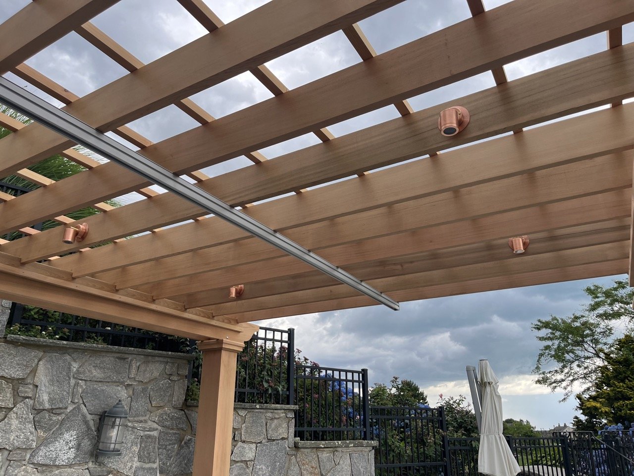 Wooden pergola with metal tracks and copper-colored light fixtures installed, outdoor setting with stone wall, black fence, trees, and cloudy sky in the background.