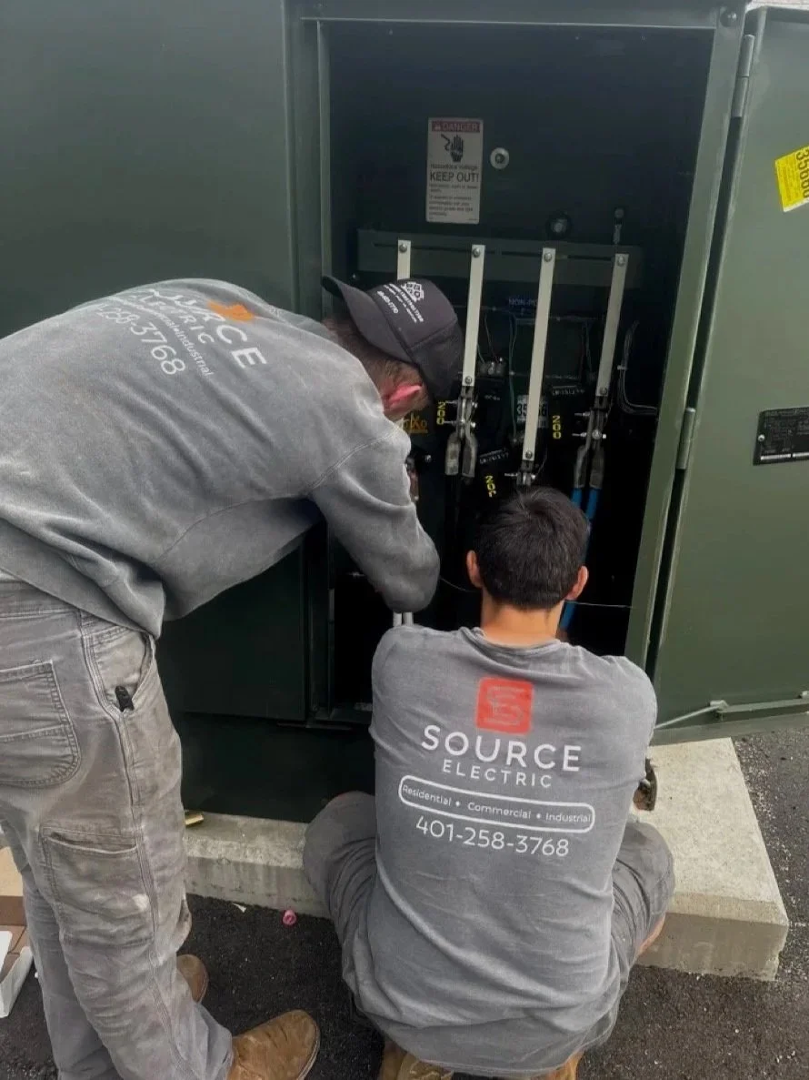 Two electrical workers are repairing or maintaining an electrical cabinet outdoors, with one person kneeling and the other standing, both working inside the cabinet.