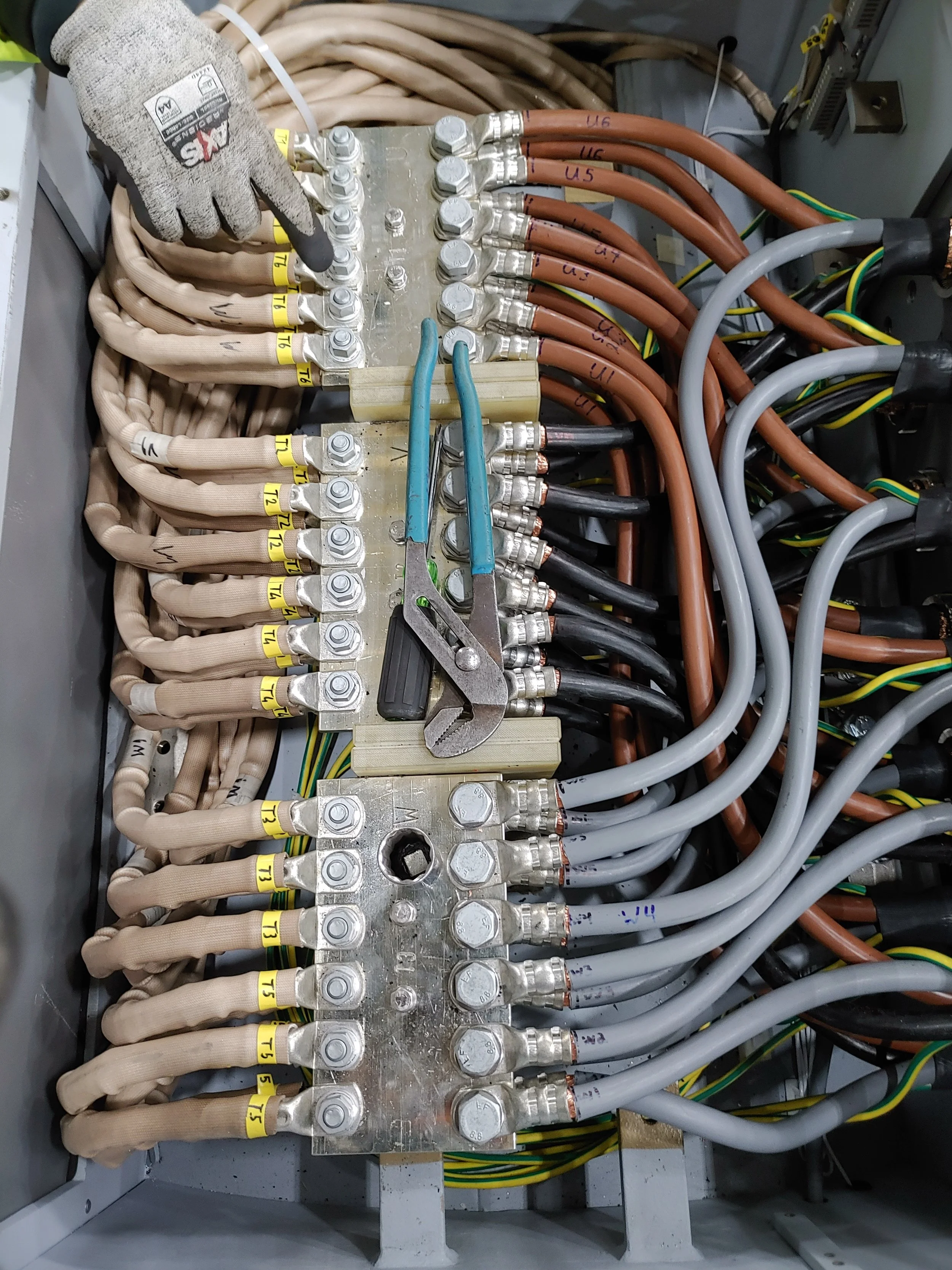 Close-up view of an electrical panel with multiple wires connected to terminal blocks. The wires are organized with color-coded insulation, including beige, black, gray, and copper-colored cables, with some labeled and secured with nuts and bolts.