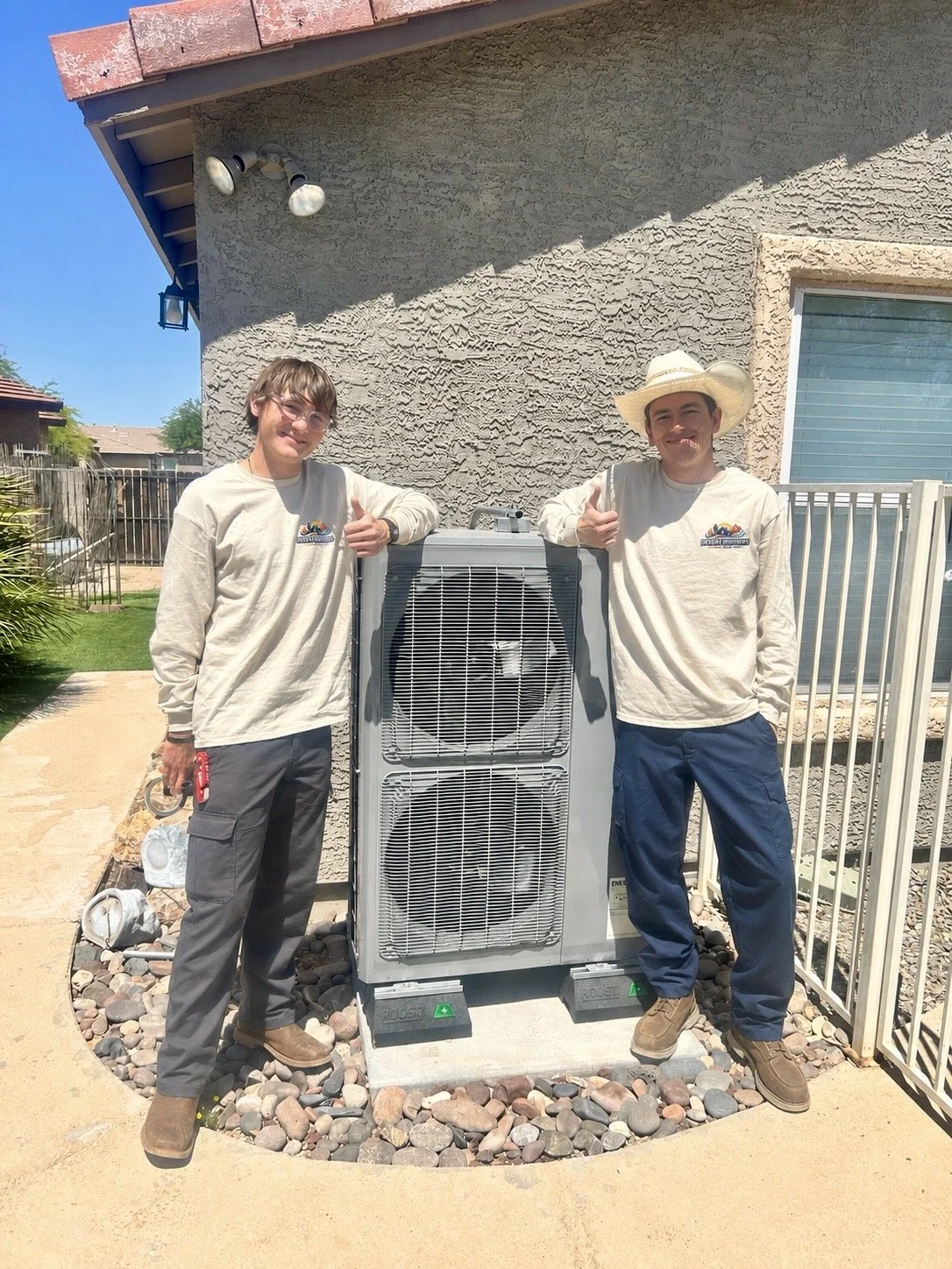 Two workers standing outside next to a large HVAC unit, giving thumbs up, with a house wall in the background.