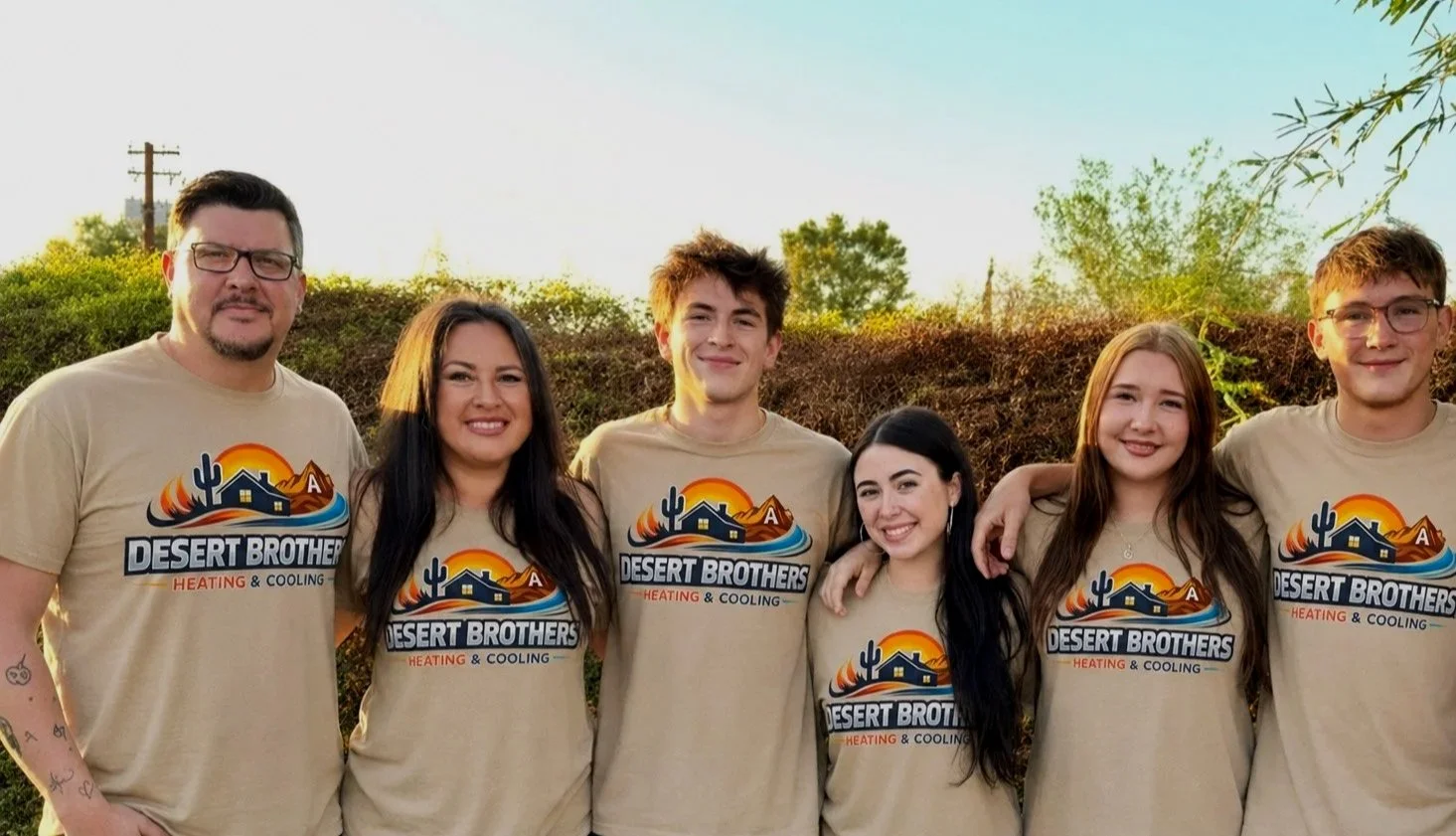 Group of six people standing outdoors with arms around each other, wearing matching beige T-shirts with a desert-themed logo that says 'Desert Brothers Heating & Cooling,' smiling during daytime.