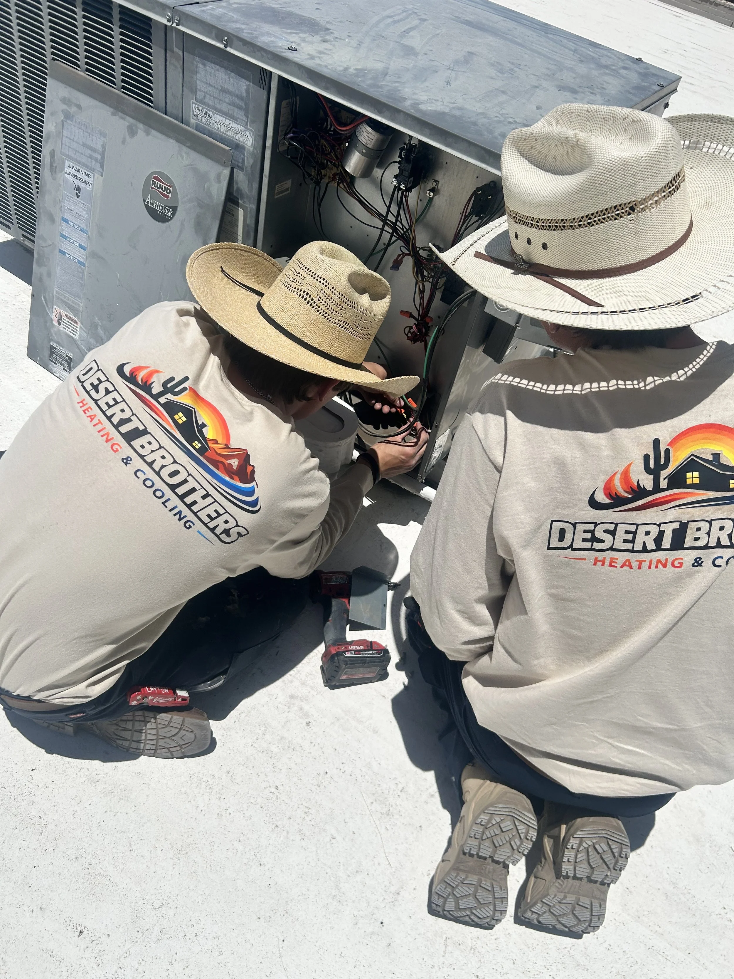 Two technicians in desert-themed shirts and straw hats working on an air conditioning unit outdoors, with tools around them.