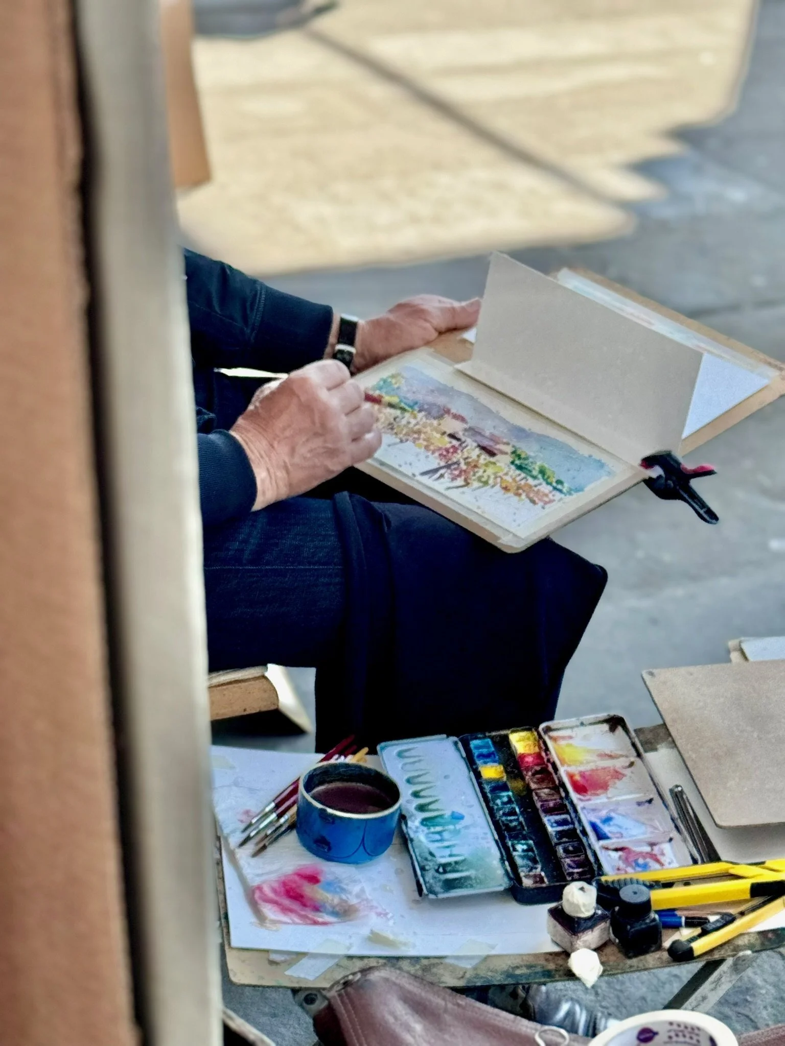 A person sitting at a table with watercolor paints and brushes, painting on paper. There are open watercolor sets, a jar with water, and various painting tools on the table.