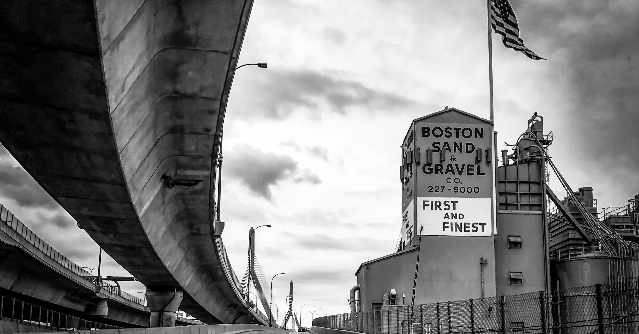 Black and white photograph of an industrial site with a marquee and a large sign for Boston Sand & Gravel, showing the company's name, phone number, and slogan 'First and Finest,' under an overcast sky with a bridge structure overhead.