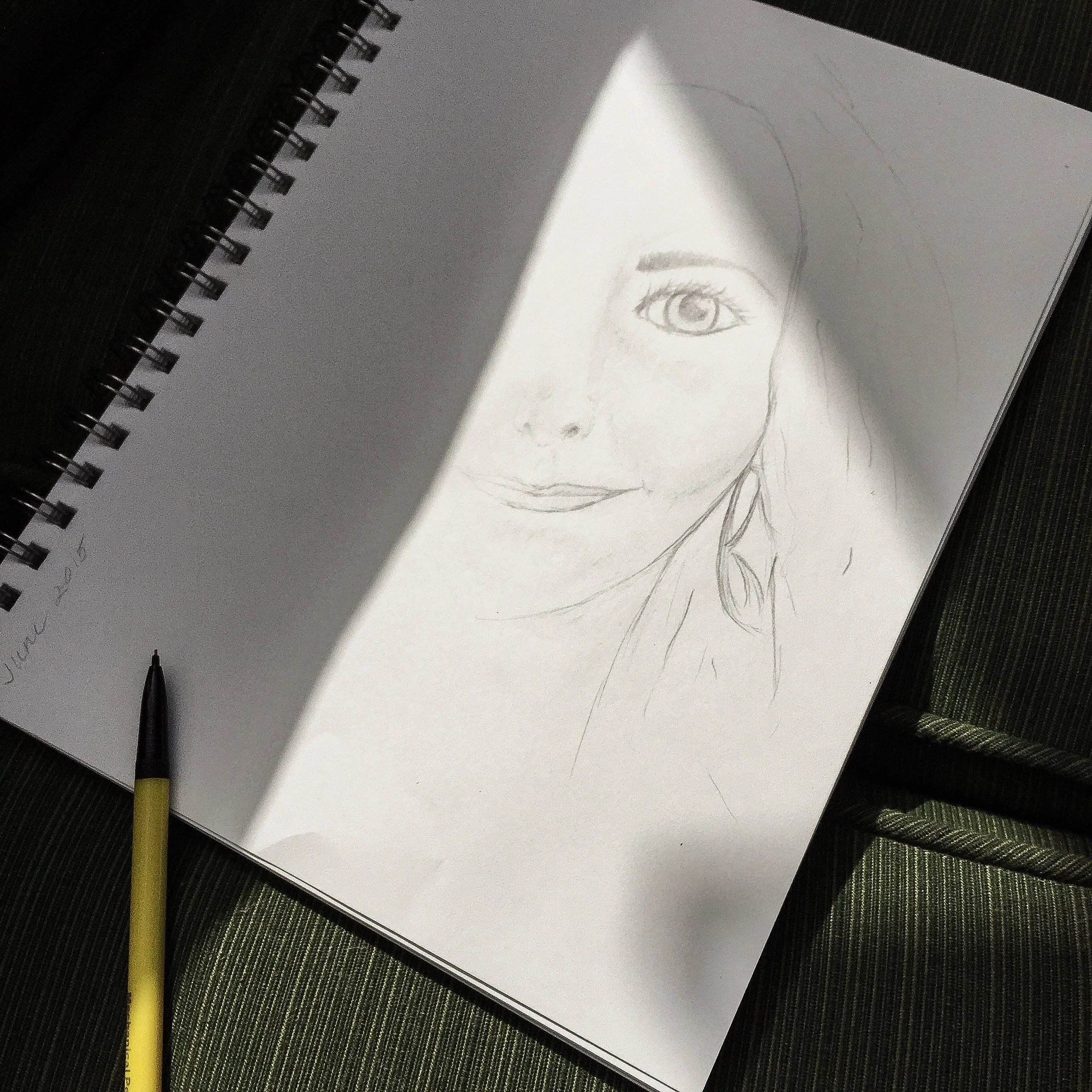 A spiral-bound notebook with a graphite pencil and a black pen resting on it, on a dark green textured surface. The notebook is open to a page with a partially drawn sketch of a woman's face, focusing on the eye, eyebrow, and part of the nose and mou