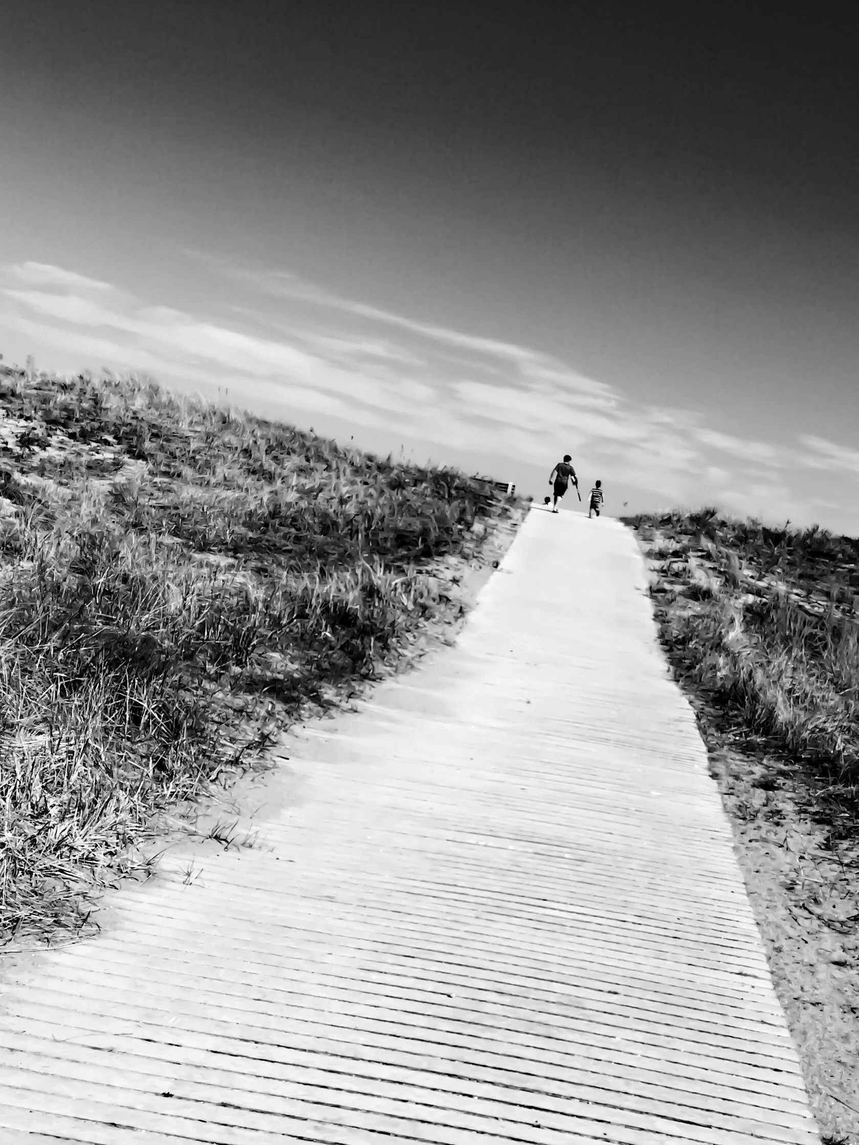 A black-and-white photograph of a wooden boardwalk path in a natural outdoor setting, with grassy areas on both sides and a person walking with a dog in the distance under a partly cloudy sky.