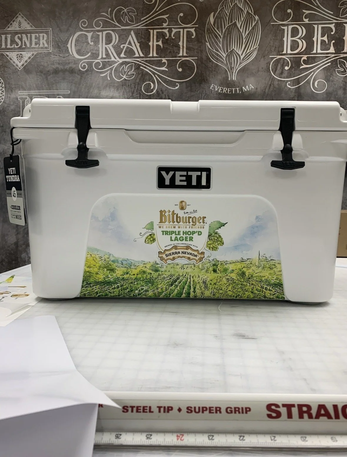 White YETI cooler with a landscape label featuring green grapevines and a mountainous backdrop, placed on a table in front of a dark wall with chalkboard-style brewery artwork.
