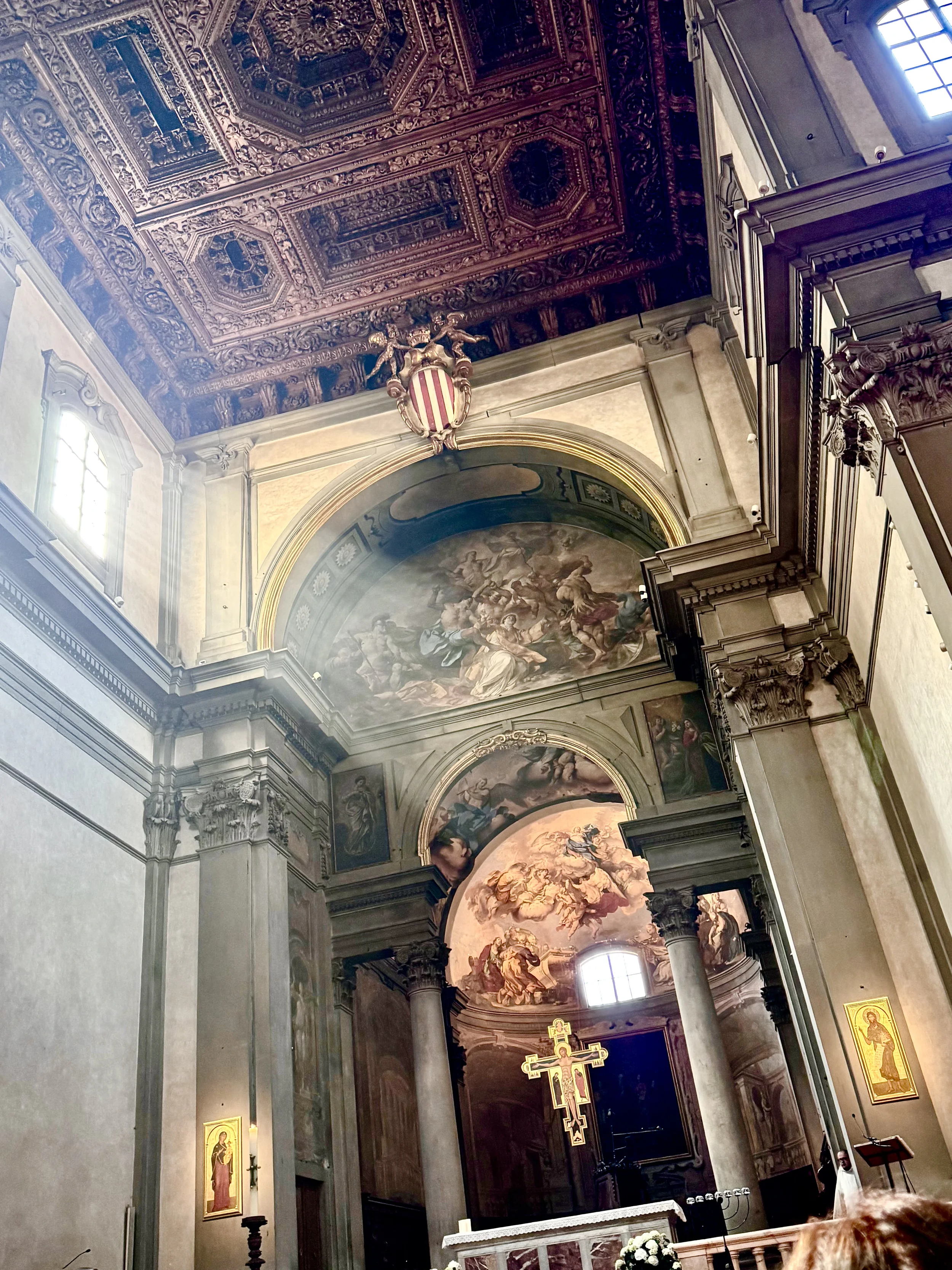 Interior of a church with ornate architectural details, including columns, arches, and a detailed ceiling. Religious artwork and icons are visible on the walls, and a large decorative cross is hanging in front of an altar.