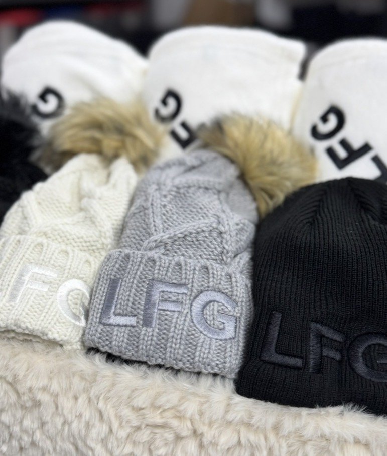 Three knit winter hats with embroidered initials LFG, LF, and LEO, in cream, gray, and black, displayed on a fluffy surface with white socks in the background.
