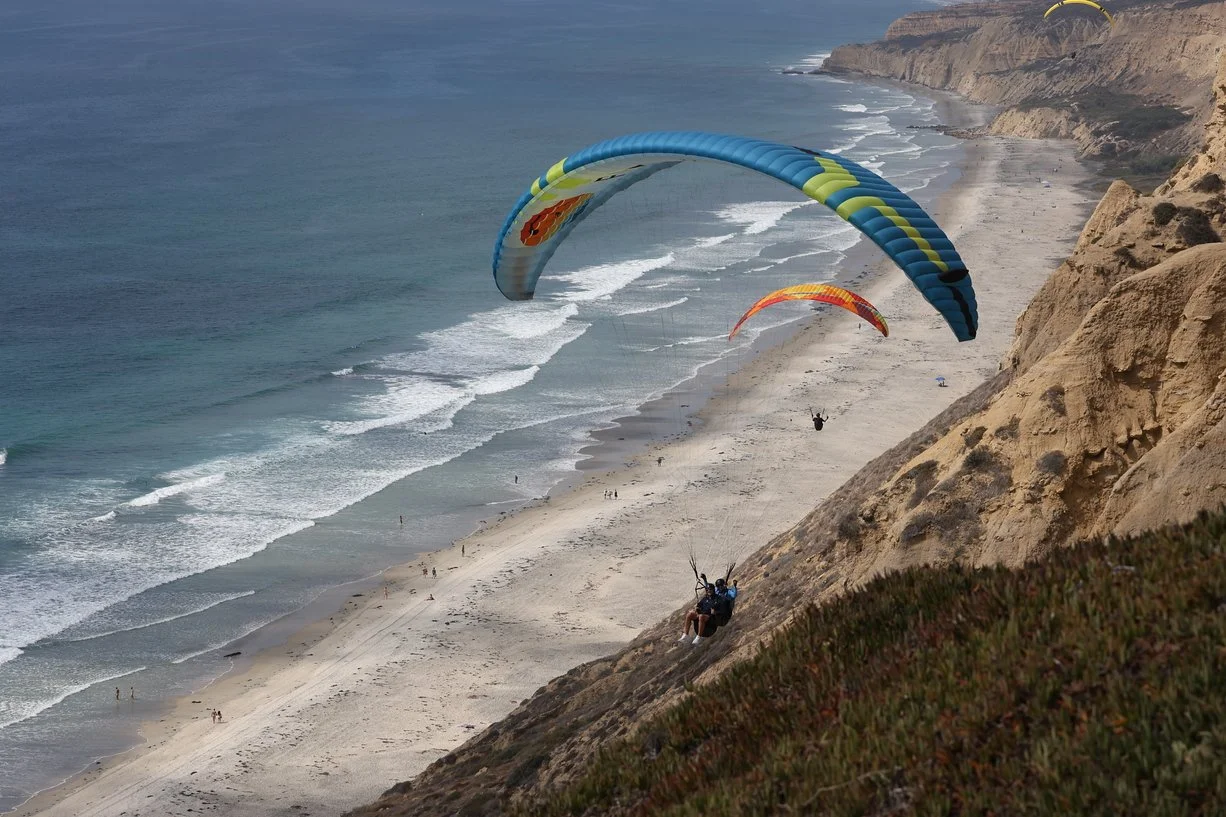 Two people paragliding over a sandy beach with cliffs on one side and the ocean on the other.