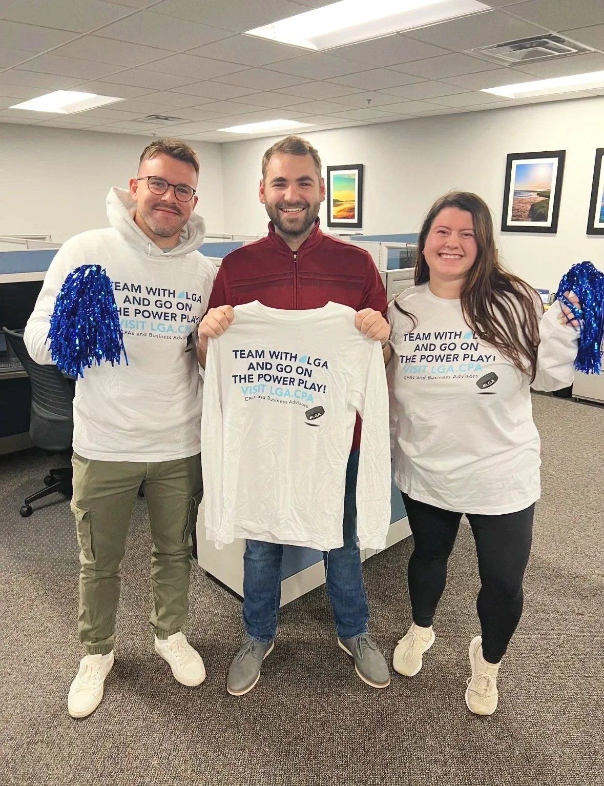 Three smiling people standing in an office, holding T-shirts and pom-poms. The T-shirts have a message about teamwork and a website. The woman on the right and the man on the left are wearing white shirts with blue text. The man in the middle is wear