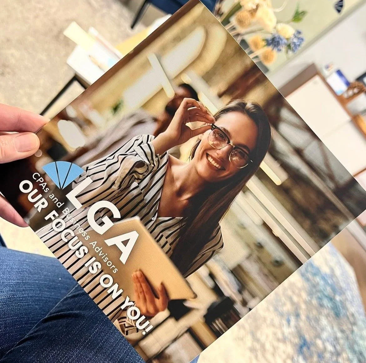 A woman with glasses smiling while holding a tablet, sitting in an indoor space with bookshelves and decor in the background.