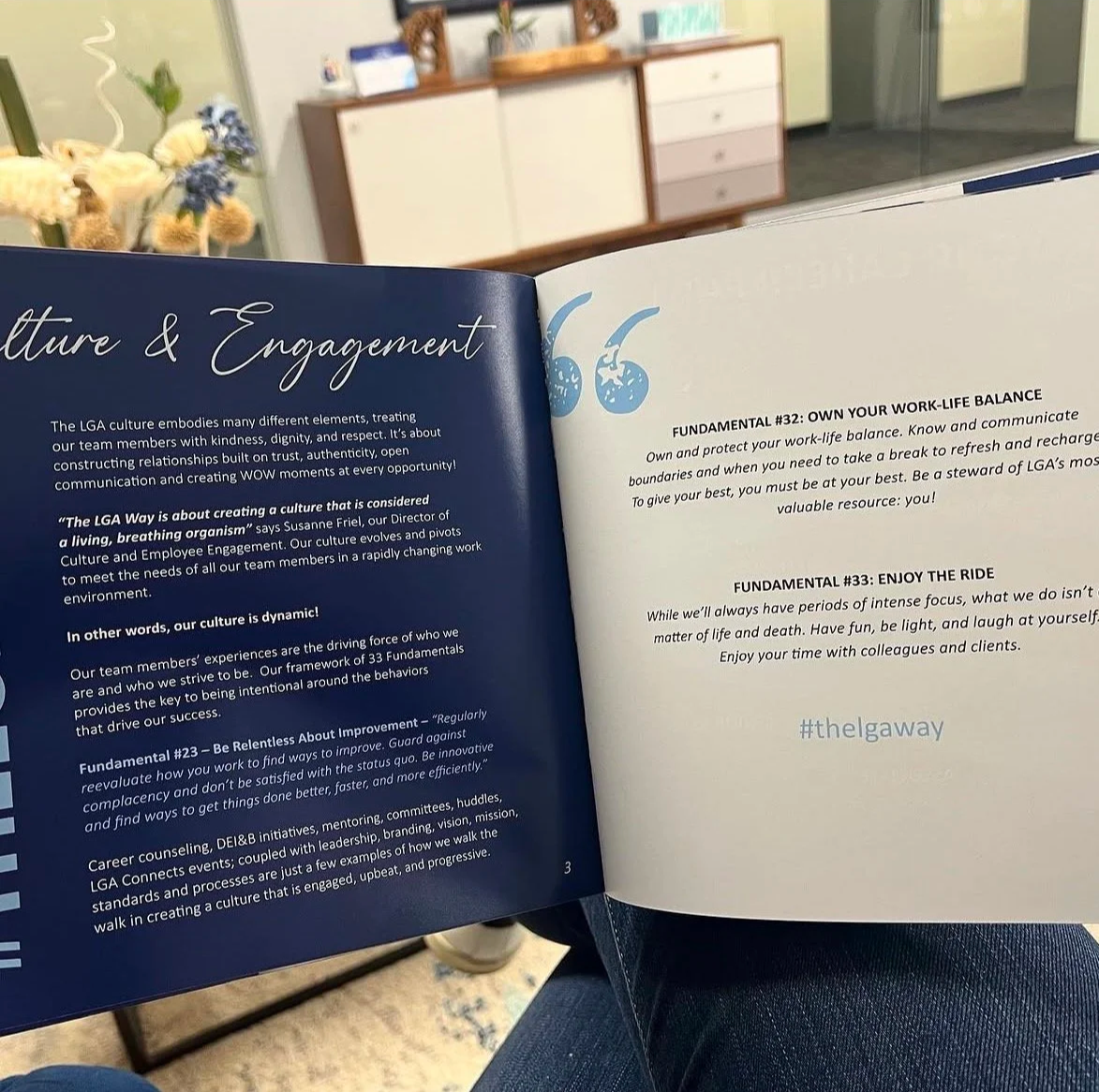 Open booklet on a person's lap with text about culture and engagement. Background shows a floral arrangement and a cabinet in room.
