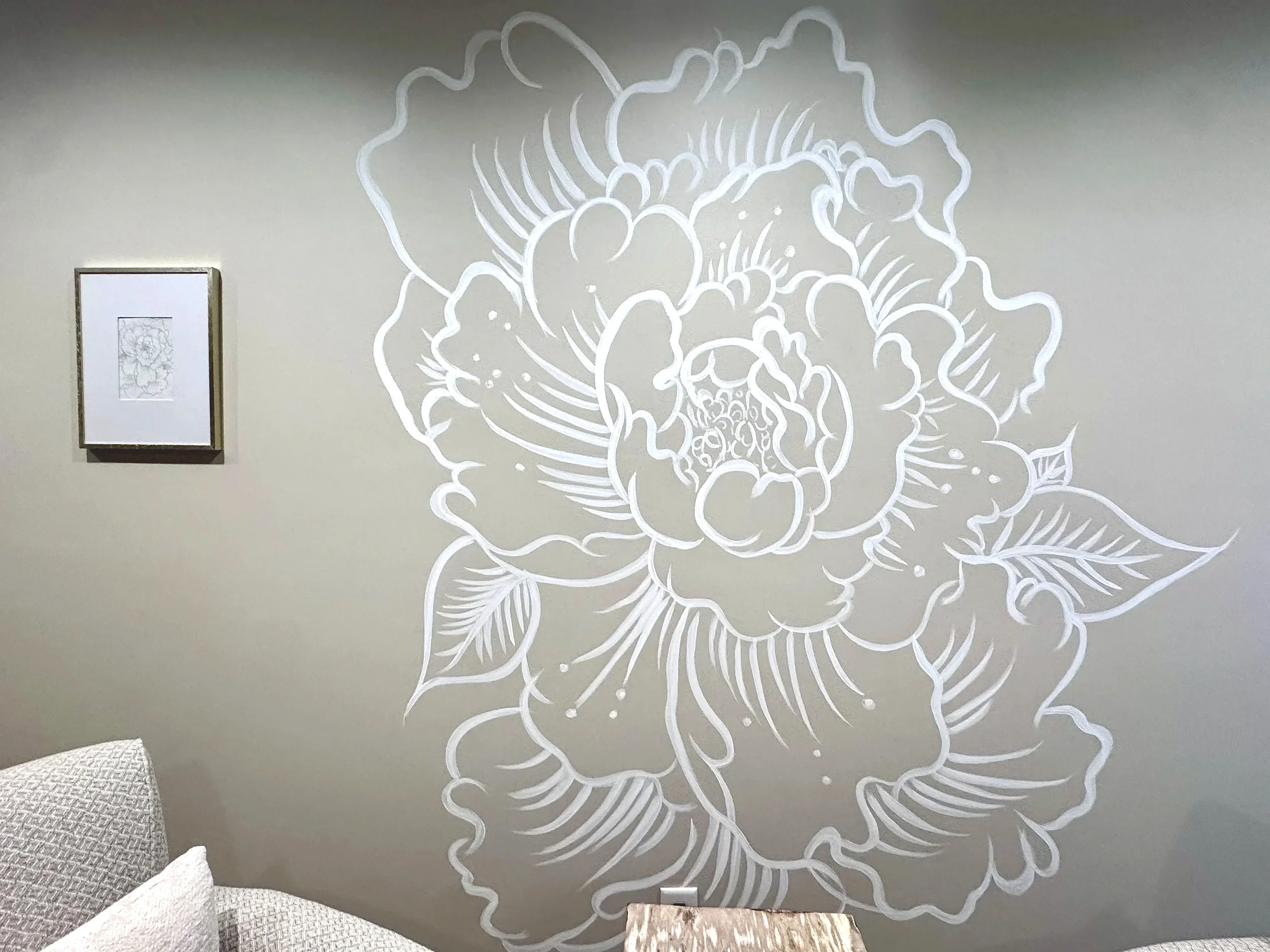 White floral line art mural of a large peony flower on a gray wall, with framed artwork and a textured gray cushion in the foreground.