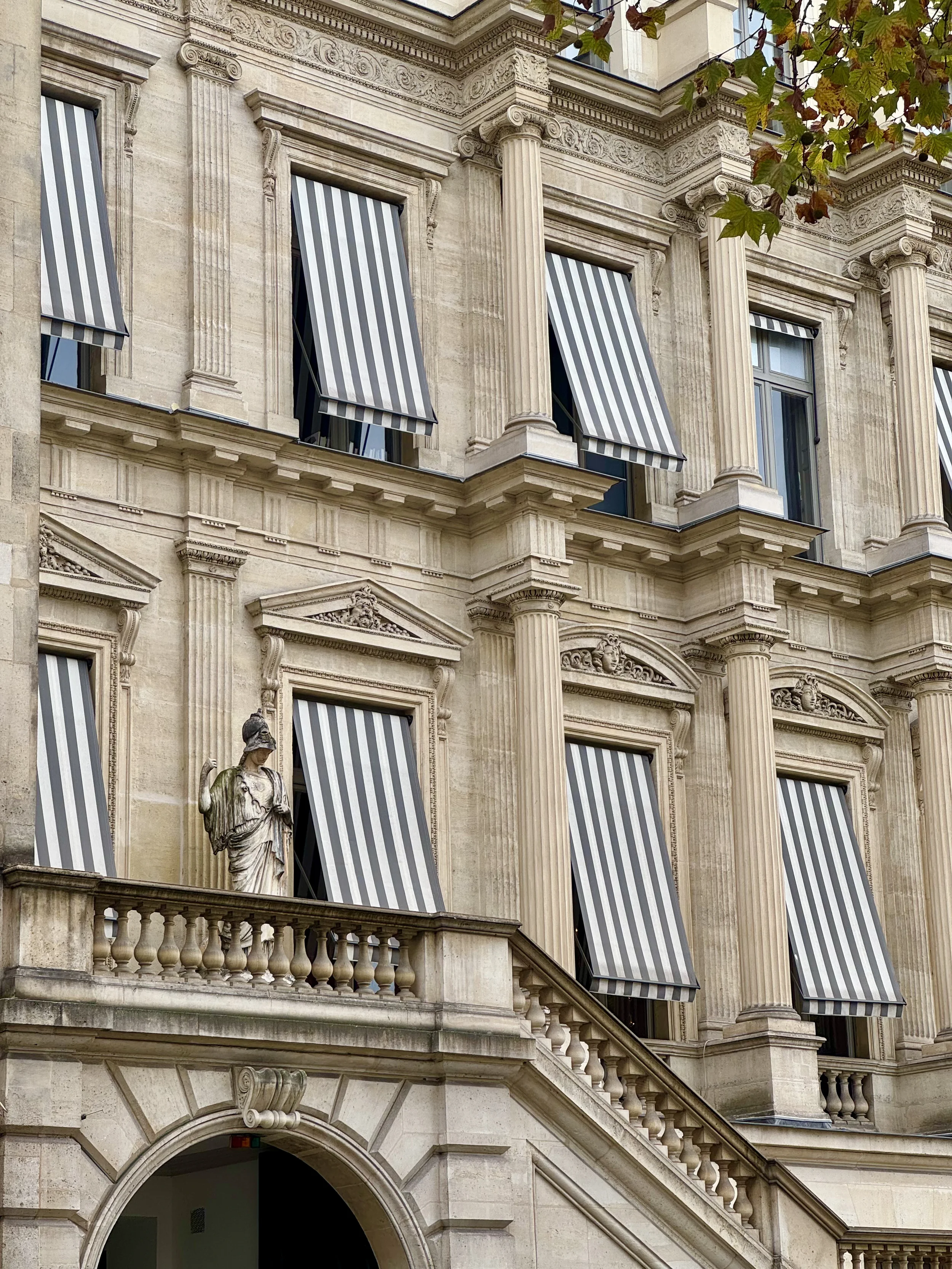Close-up of a classical-style building exterior with ornate stone details, Roman columns, and black-and-white striped awnings over the windows, with some autumn leaves visible at the top.
