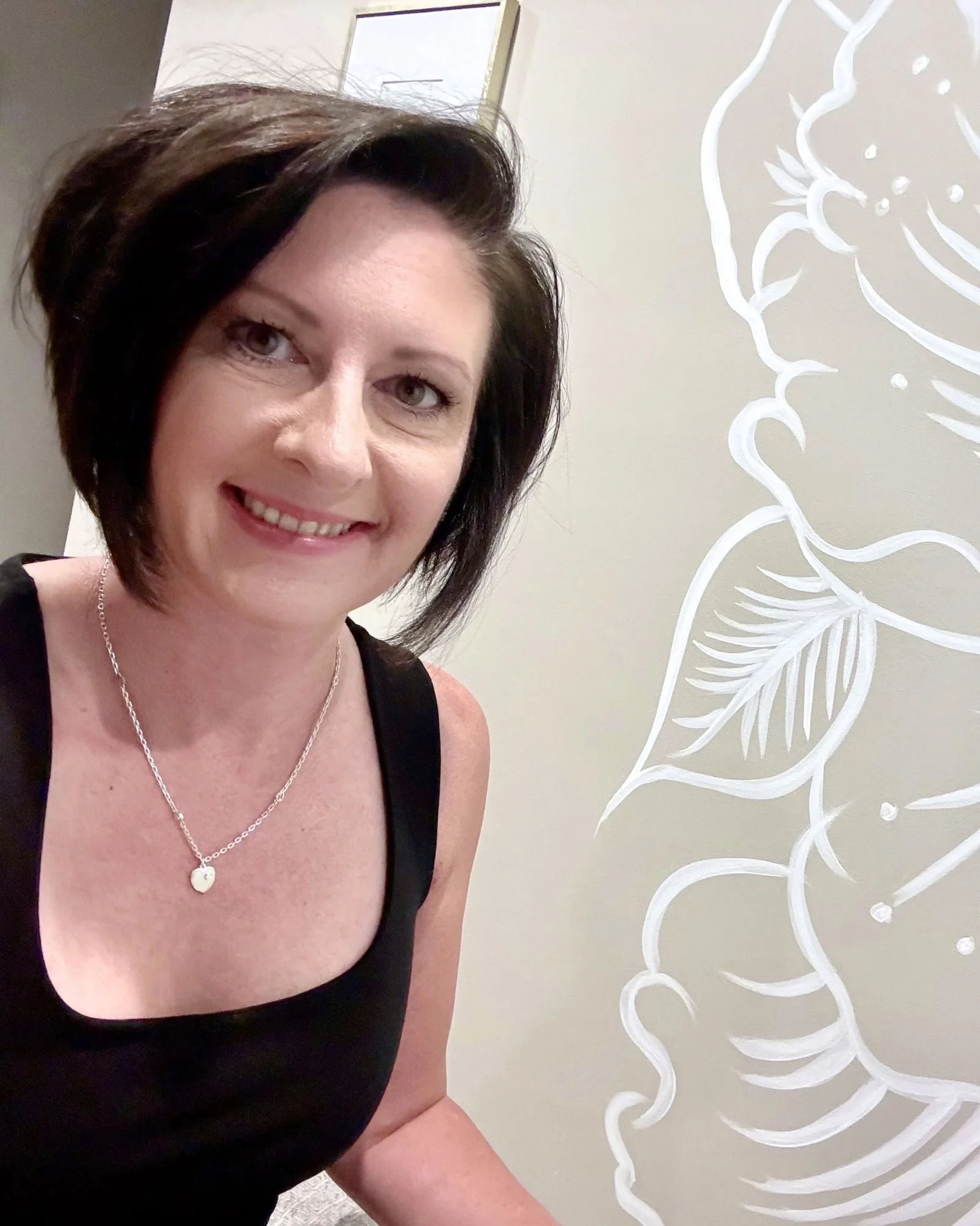 Close-up of a woman smiling, wearing a black tank top and a heart-shaped necklace. Part of her face and upper body are visible, with a decorative white floral wall art next to her.