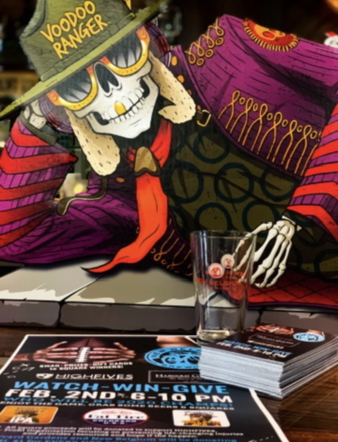 Colorful illustration of a skeleton wearing sunglasses, a hat with 'Voodoo Ranger' written on it, and a purple and red outfit, sitting with a glass nearby, at a promotional booth.