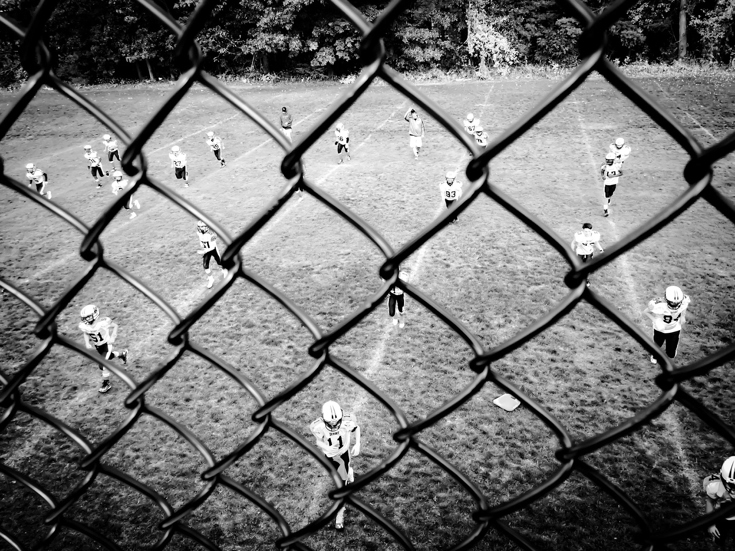 Black and white photo of football players on a field, viewed through a chain-link fence, with players wearing helmets and uniforms with numbers.