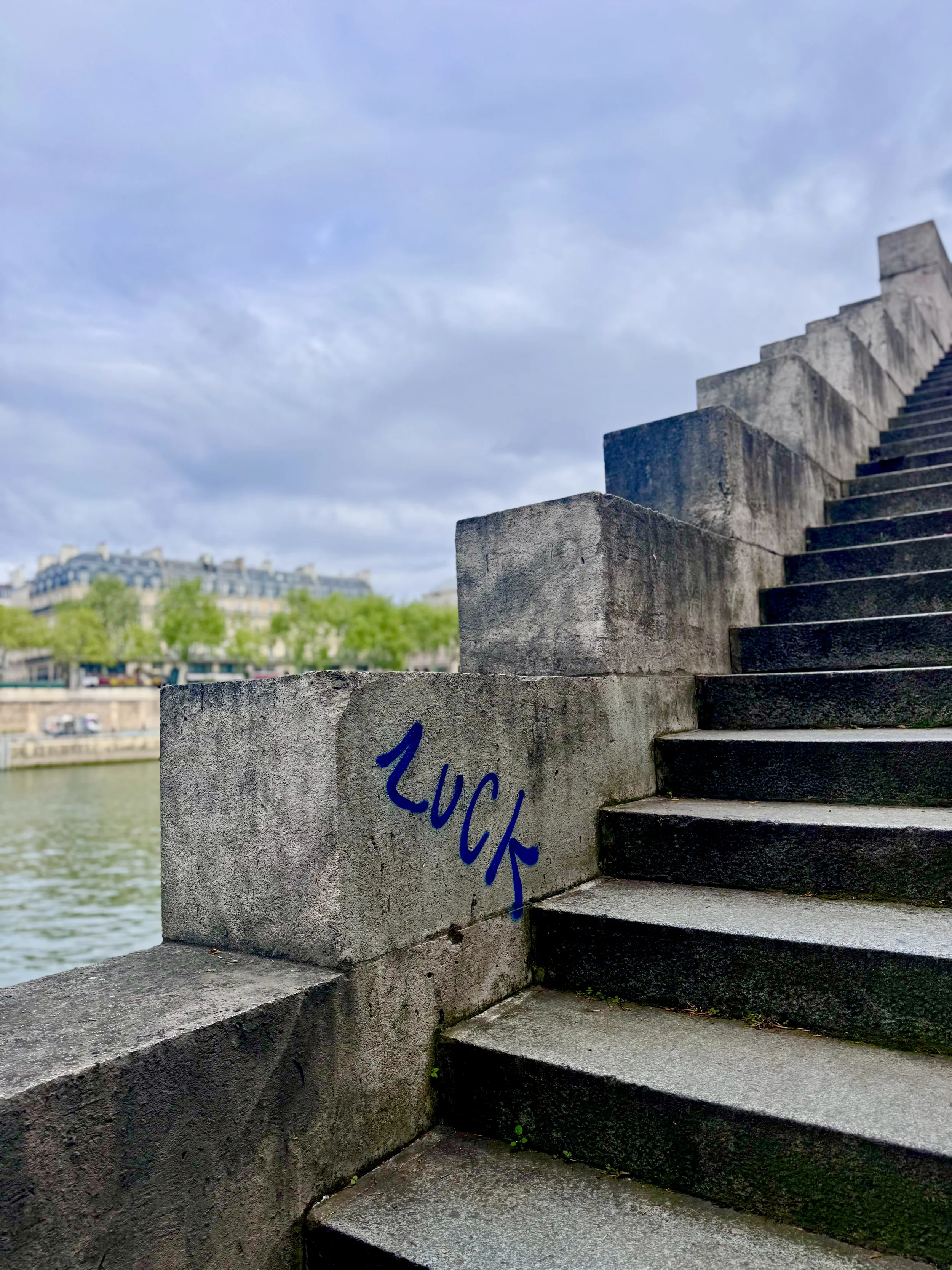 Concrete stairs with the word 'Lucky' written in blue on the side, overlooking a river with a cityscape and buildings in the background under a cloudy sky.