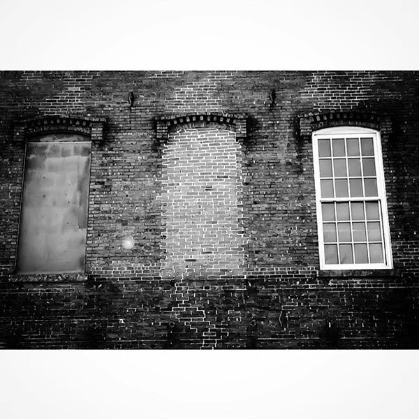 A black and white photo of a brick building's exterior wall with three windows, one boarded up, one with a window, and one without.