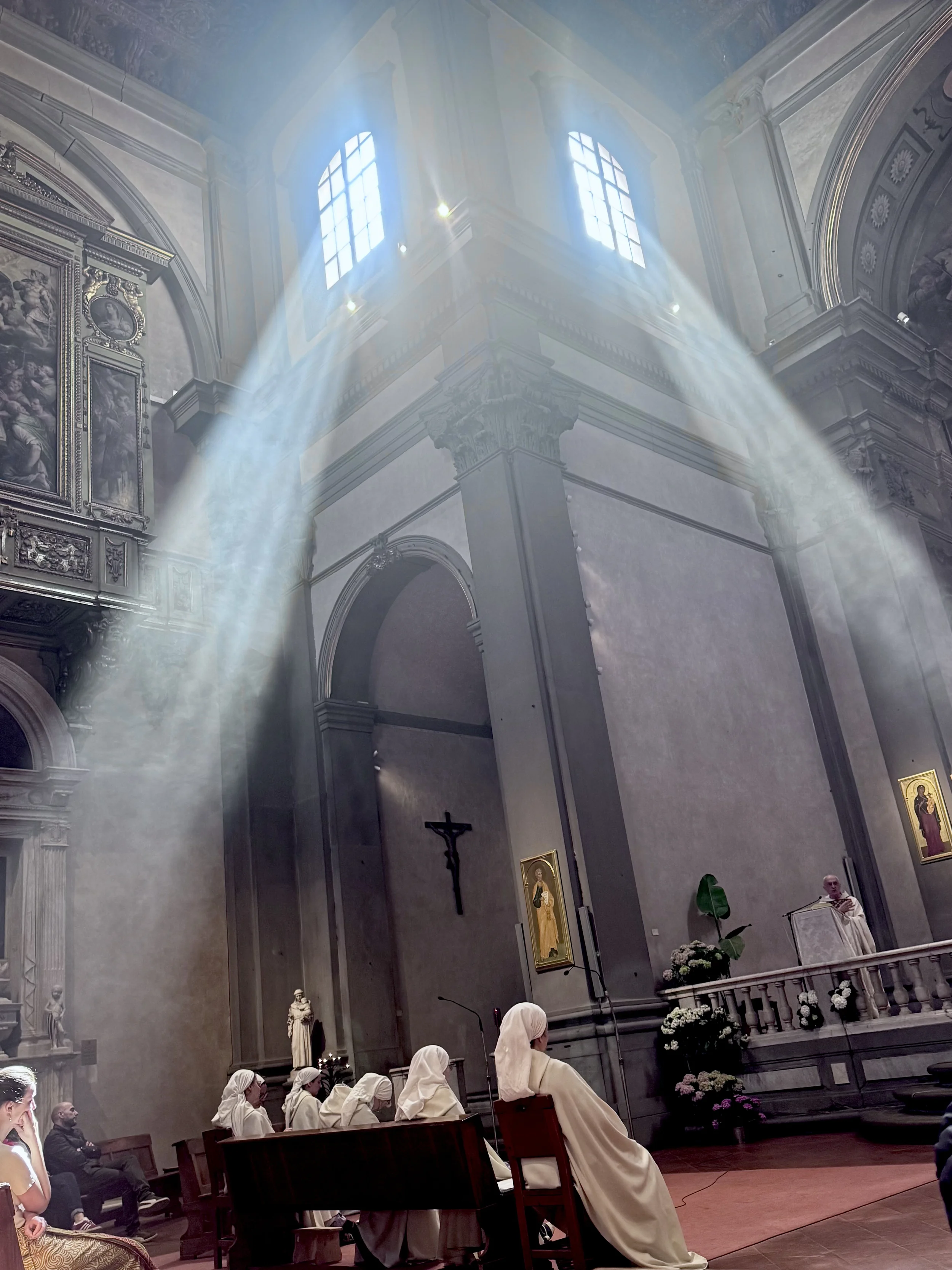 Inside a church with sunlight streaming through stained glass windows. People, including nuns, are seated in pews, facing the altar where a priest is speaking. Religious paintings and statues adorn the interior walls.