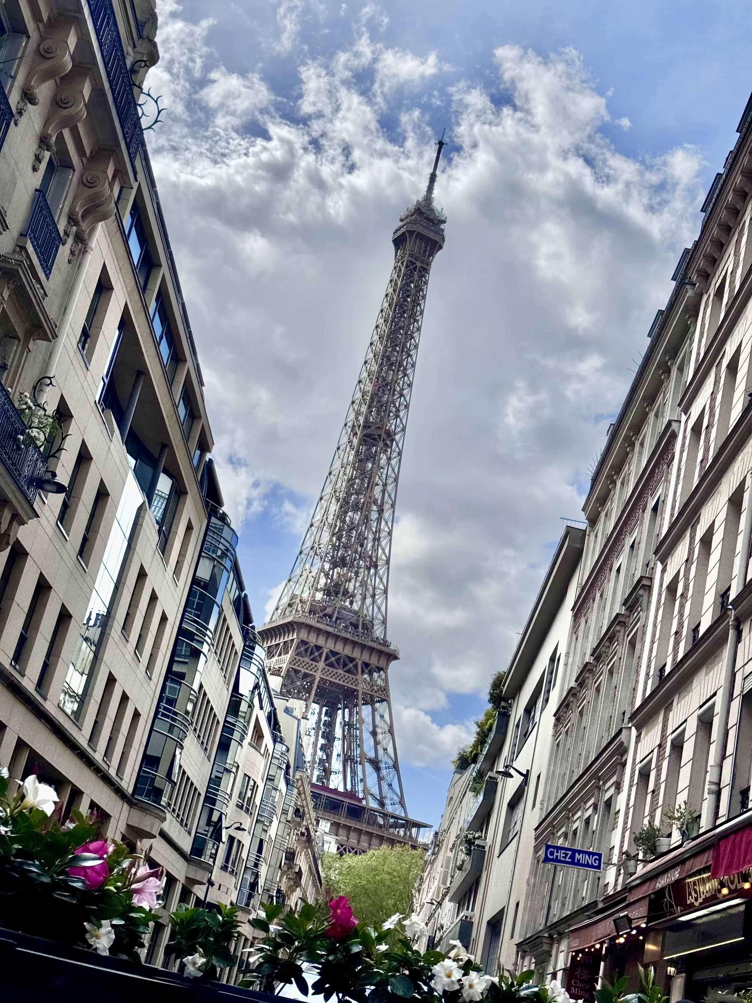 View of the Eiffel Tower from a narrow street in Paris, flanked by multi-story buildings with balconies. The sky is partly cloudy, and there are flowers in the foreground.