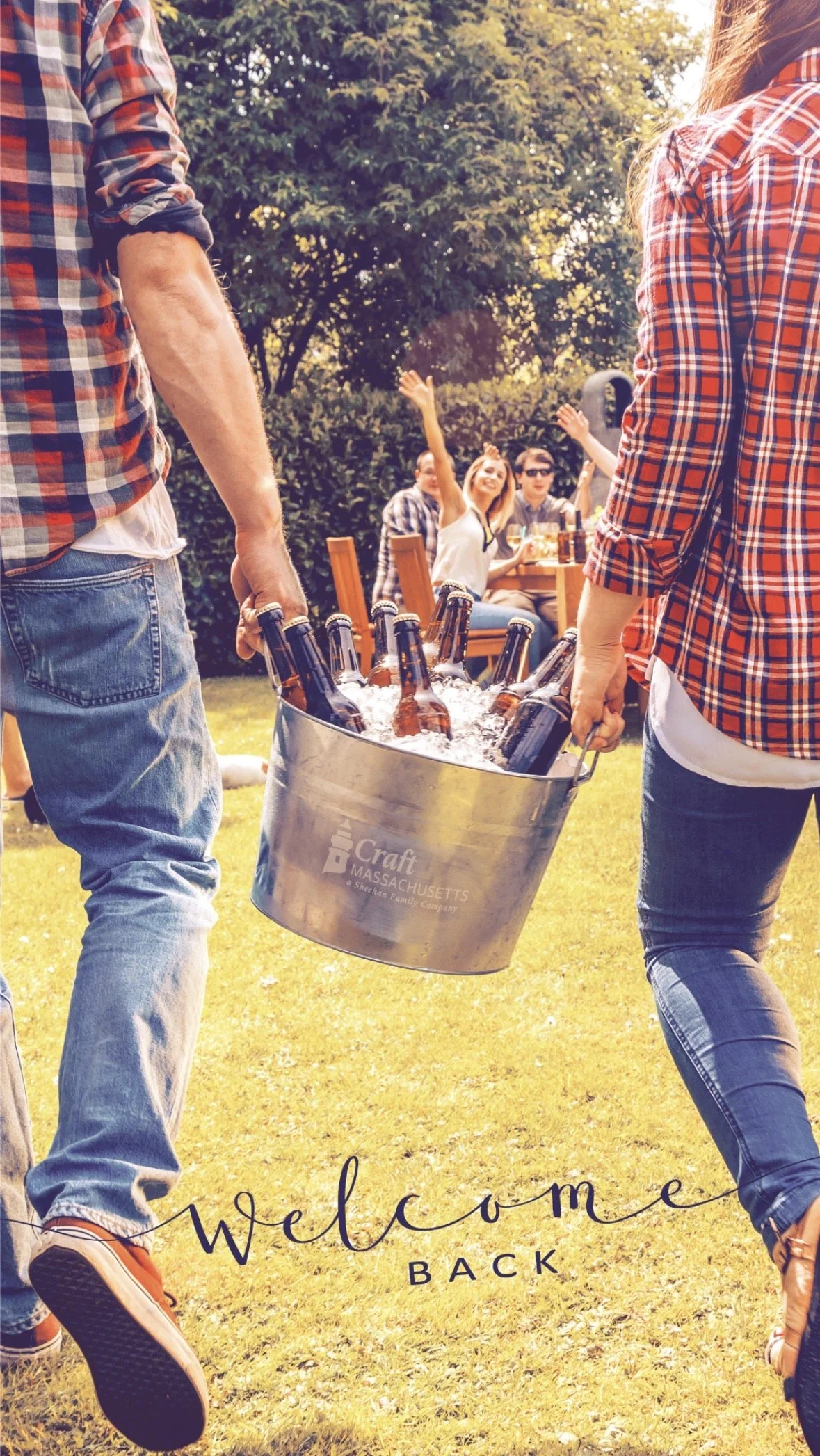 People carrying a metal bucket filled with bottles of beer at an outdoor gathering, with a group of friends sitting at a table in the background, enjoying a sunny day.