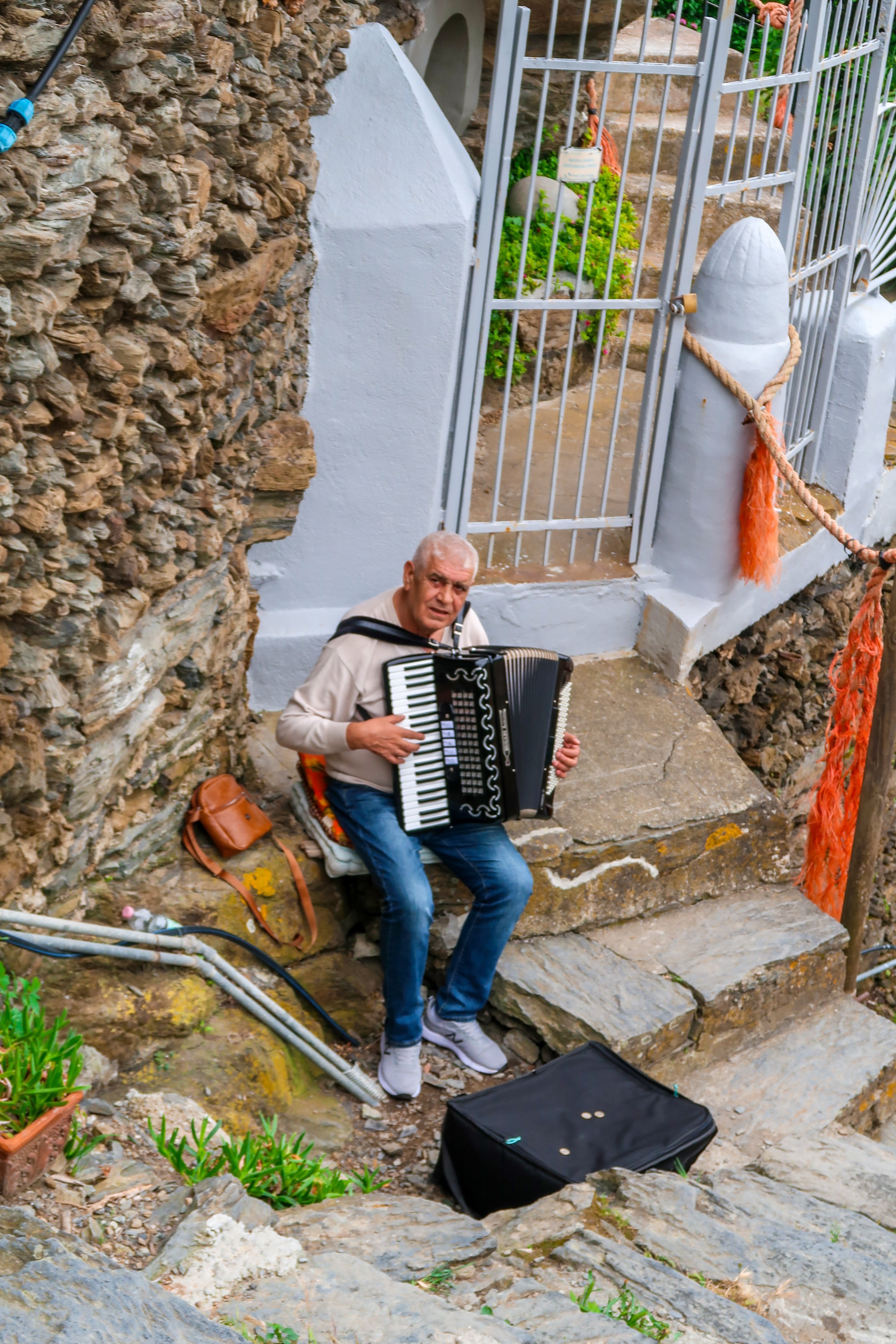 An elderly man playing an accordion while sitting on stone steps outdoors near a stone wall and a metal gate, with some personal belongings nearby, including a brown bag and a black instrument case.