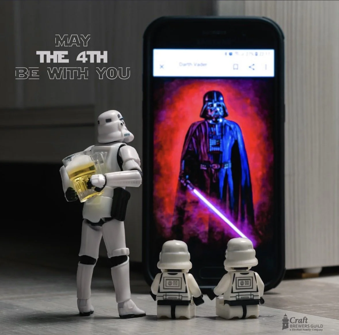 Star Wars themed figurines of a stormtrooper and two droids, with a smartphone screen displaying Darth Vader in the background, and the text "May the 4th be with you" nearby.