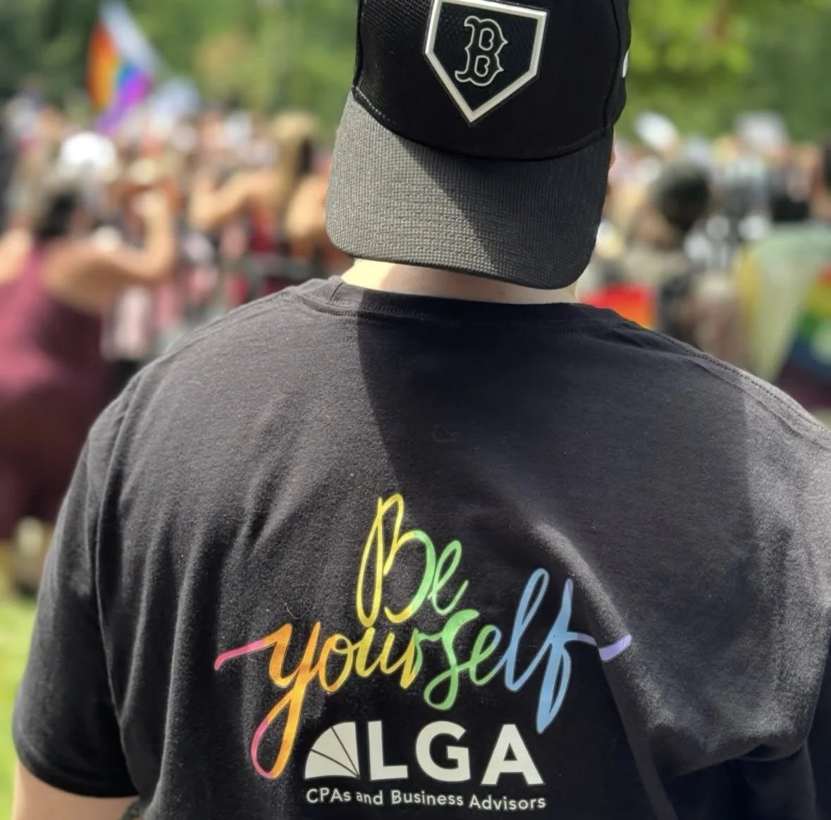 Back view of a person wearing a black baseball cap with a white logo and a black T-shirt with colorful text that says "Be yourself" and a logo below it for 'LGA CPAs and Business Advisors'. In the blurred background, people are gathered outdoors.