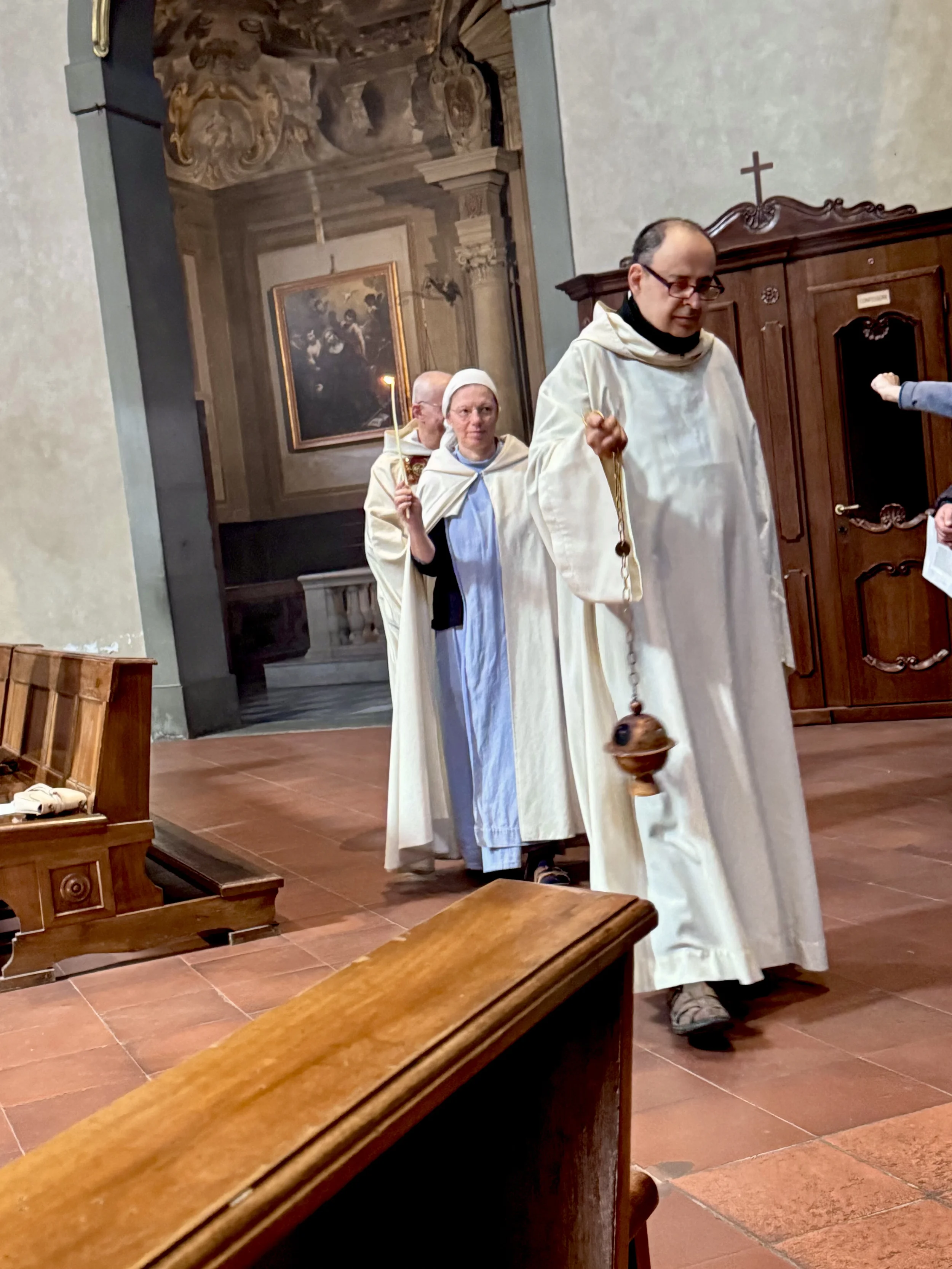 Three priests or clergy members dressed in white robes participate in a religious procession inside a church. One carries a small bell, and another holds a lit candle. The church interior features ornate artwork, religious paintings, and wooden furni