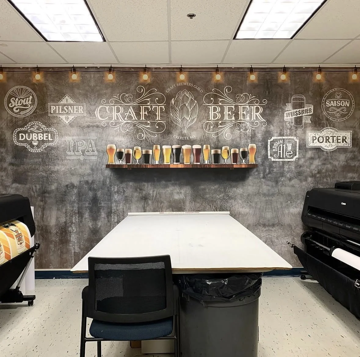 Craft beer tasting room with a wall display of various beer styles and a shelf of beer glasses, a white table, a black chair, and printing equipment.