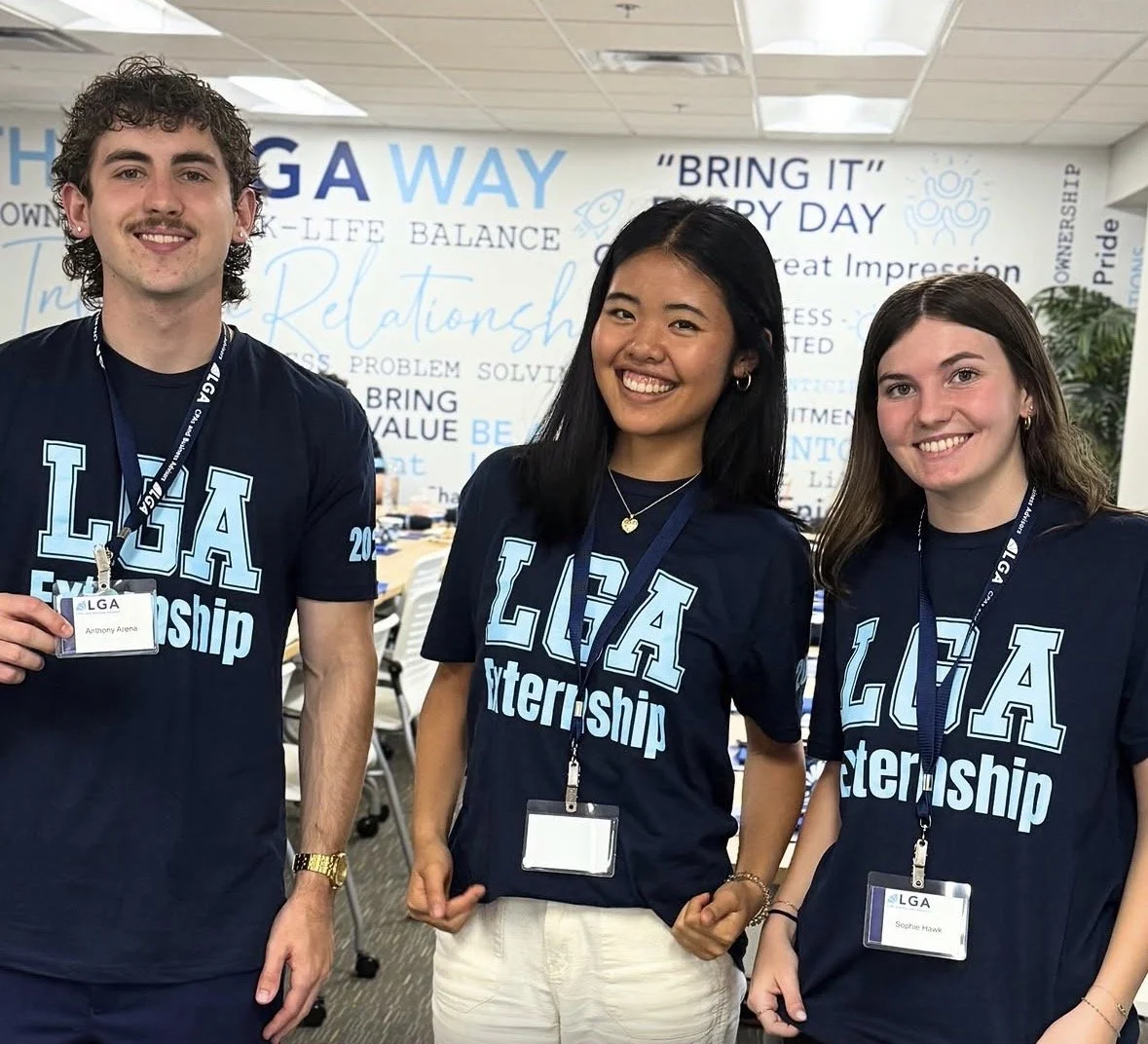 Three young people wearing navy blue LGA Internship T-shirts and name badges, standing and smiling in an office environment with motivational words on the wall behind them.