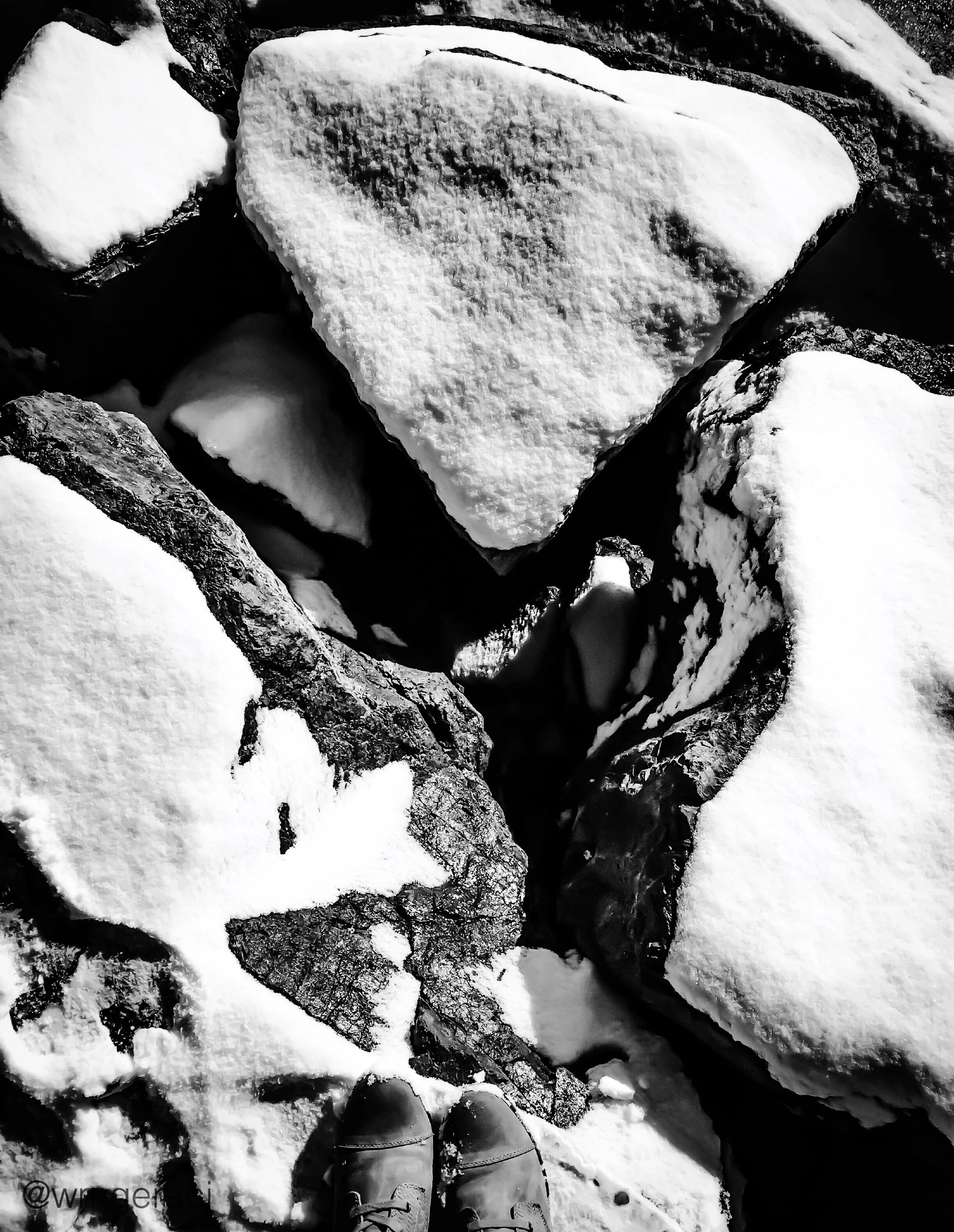 Black and white photo taken from the perspective of a person standing on snow-covered ground, wearing winter boots. The person's shadow is cast on the snow. There are numerous rocks and snow patches visible on the ground, indicating a rugged outdoor 