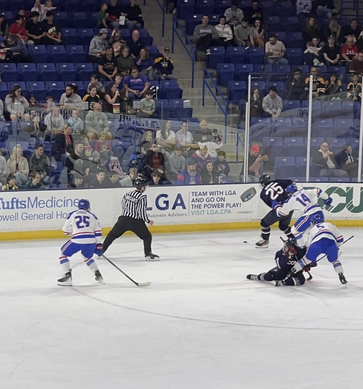 An ice hockey game with players in black and white jerseys engaged in a play, a referee watching, and spectators seated behind protective glass in the stands.