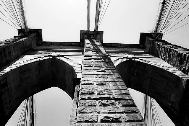 Black and white photo of the Brooklyn Bridge's stone arch and suspension cables from below, looking up.