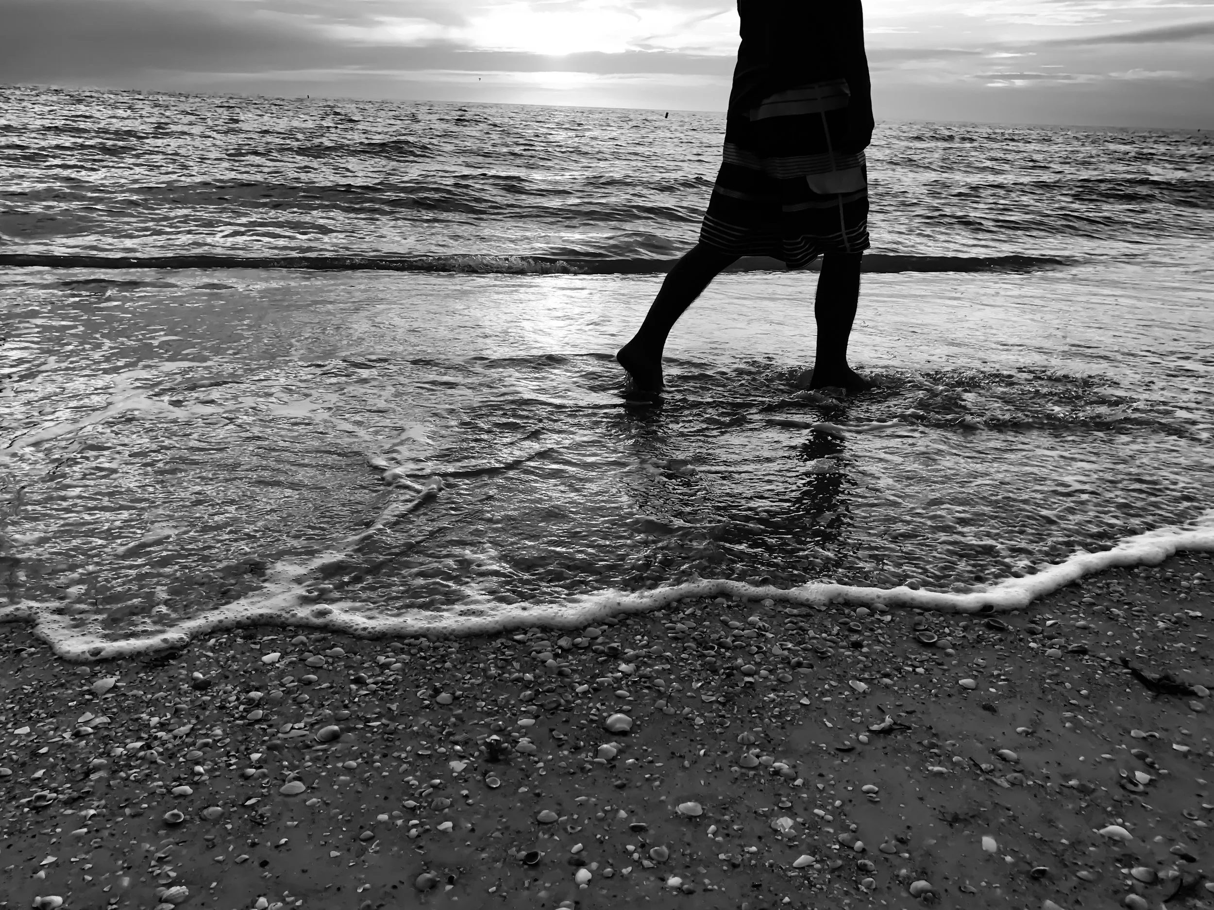 A person standing on a beach with their feet in the wet sand, facing the ocean. The person is wearing striped shorts and a dark top. The photo is black and white, with waves gently crashing on the shore and the sun setting or rising over the horizon.