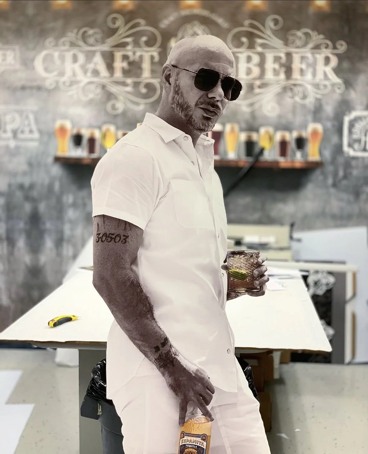 A man with sunglasses in a white shirt and pants holding a glass of a dark beverage with lime, standing in a bar or brewery with beers on a shelf and a chalkboard sign in the background.
