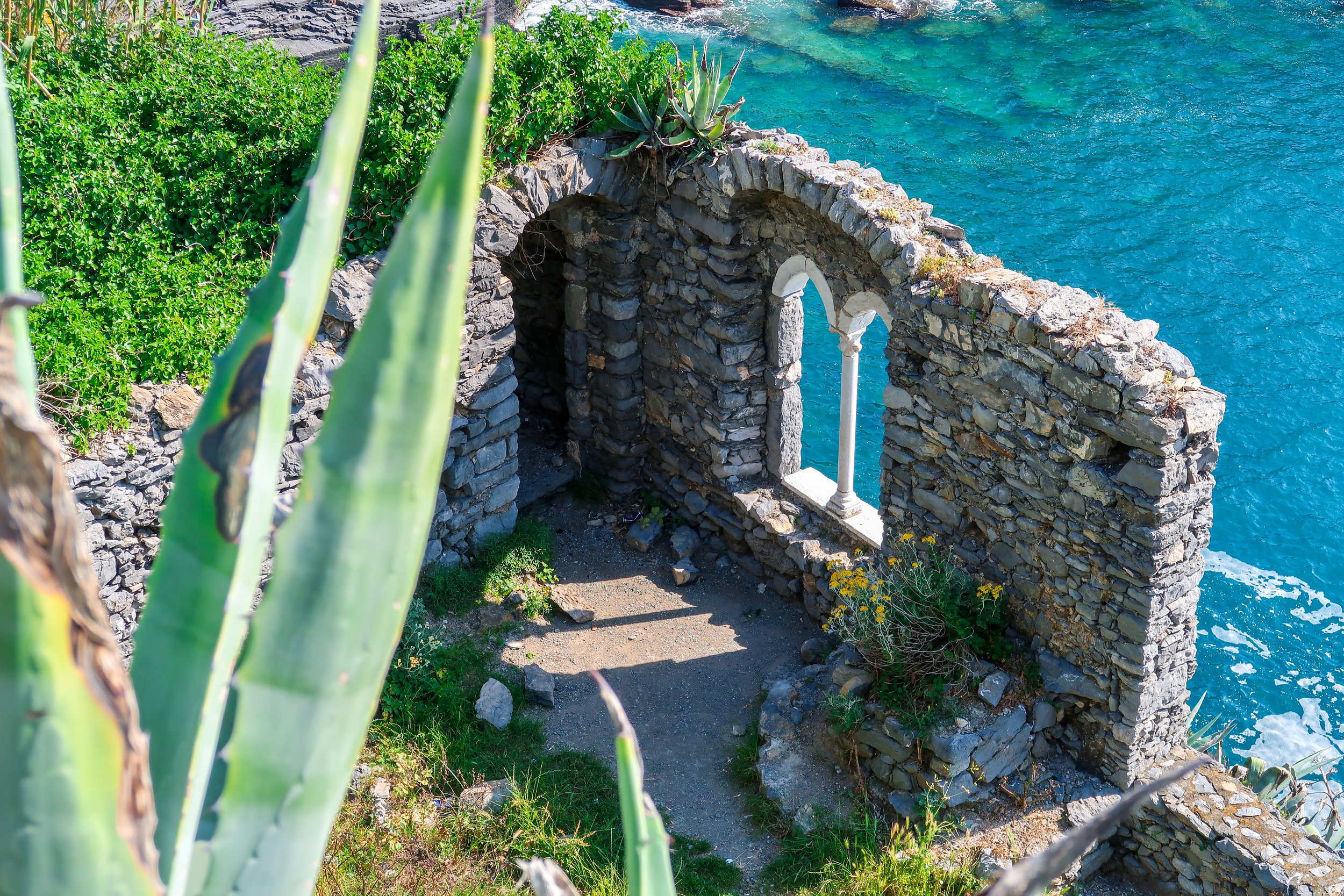View of a stone archway on a rocky cliffside overlooking the blue ocean, with green vegetation and succulent plants in the foreground.