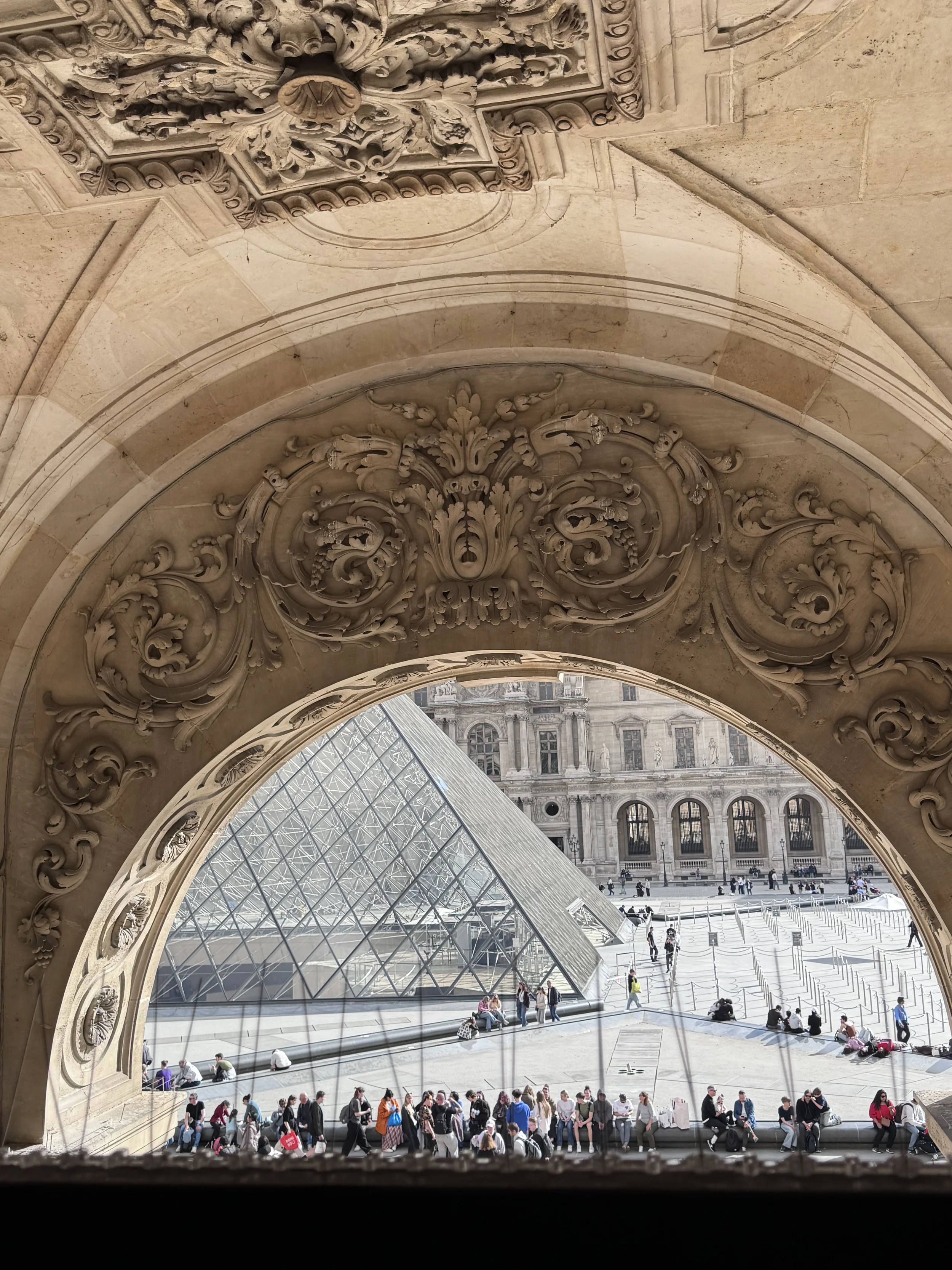 View of the courtyard of the Louvre Museum in Paris, France, seen through an ornate stone archway. The courtyard has people sitting and walking, with the Louvre's glass pyramid structure visible in the background.