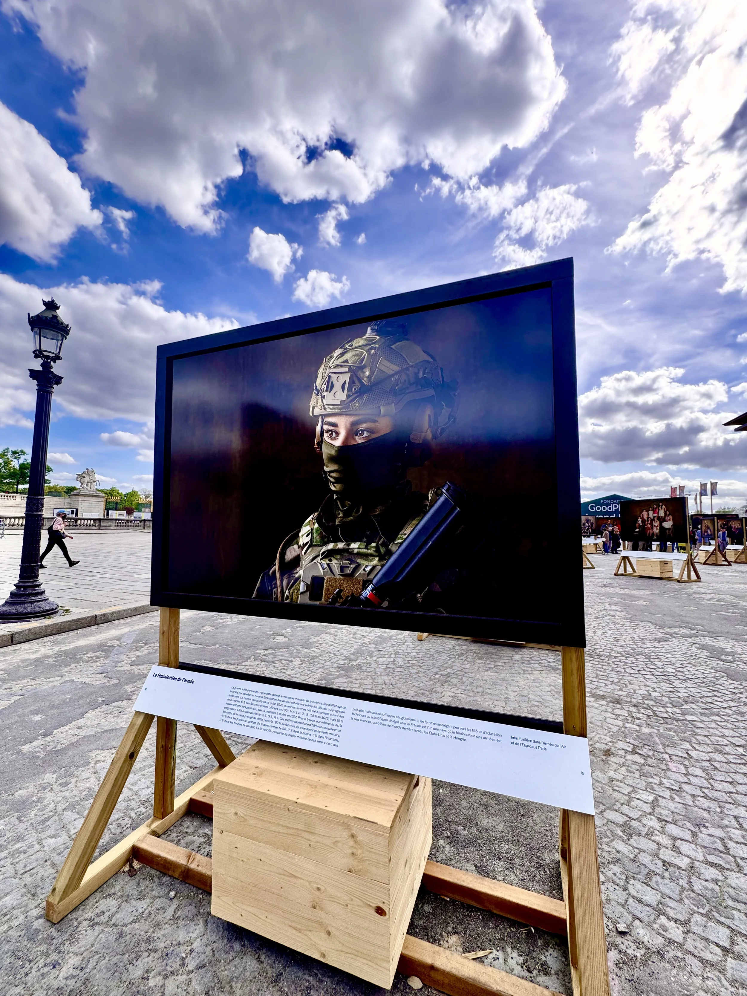 An outdoor display featuring a large photograph of a soldier wearing military gear, including a helmet and face mask, with a cloudy sky in the background. The photo is mounted on a black frame and is supported by a wooden stand with an informational 
