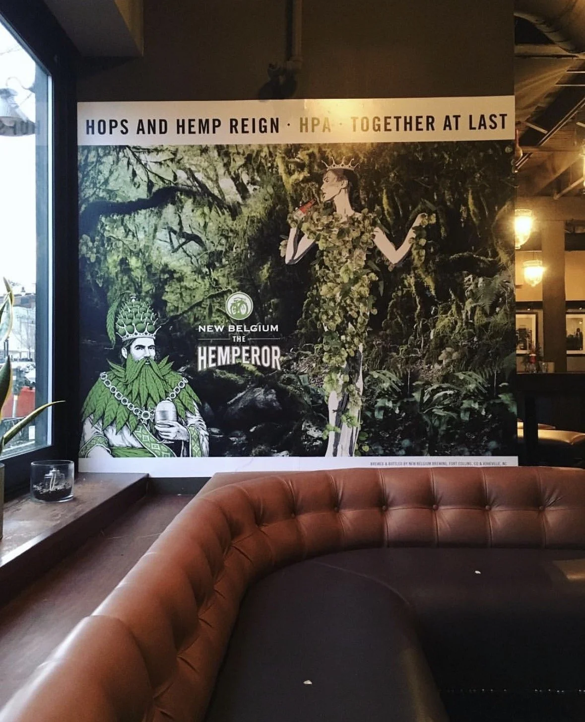A poster in a restaurant promoting a beer with a nature theme, featuring a woman in a dress made of hops and a man in a crown made of hops and hemp leaves, with the text 'Hops and Hemp Reign' and 'HPA Together at Last.'