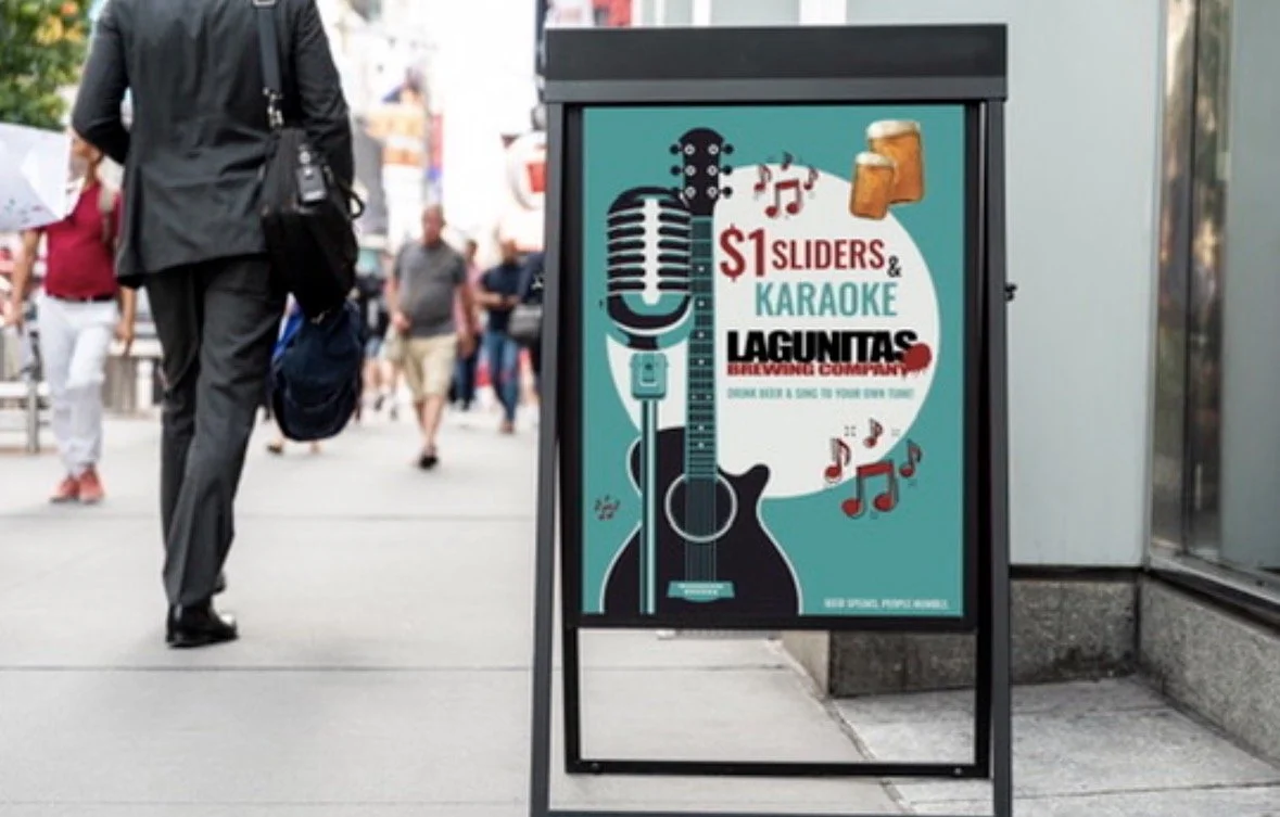 Advertising sign for Lagunitas Brewing Company promoting $1 sliders and karaoke, featuring illustrations of a microphone, guitar, and beer mugs, on a city sidewalk with pedestrians in the background.